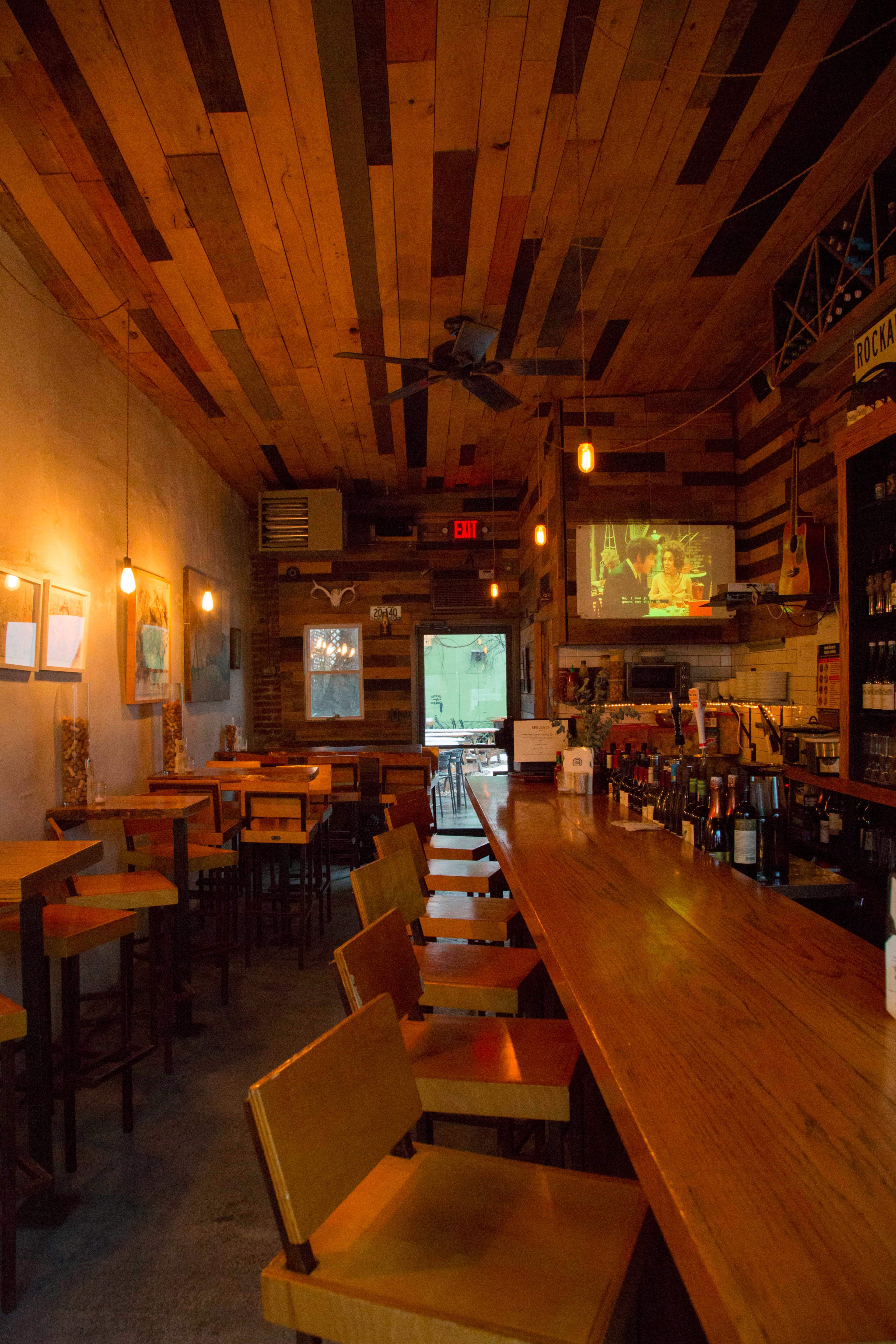An empty bar area with wooden chairs and a long counter, featuring warm lighting and exposed wooden beams on the ceiling.