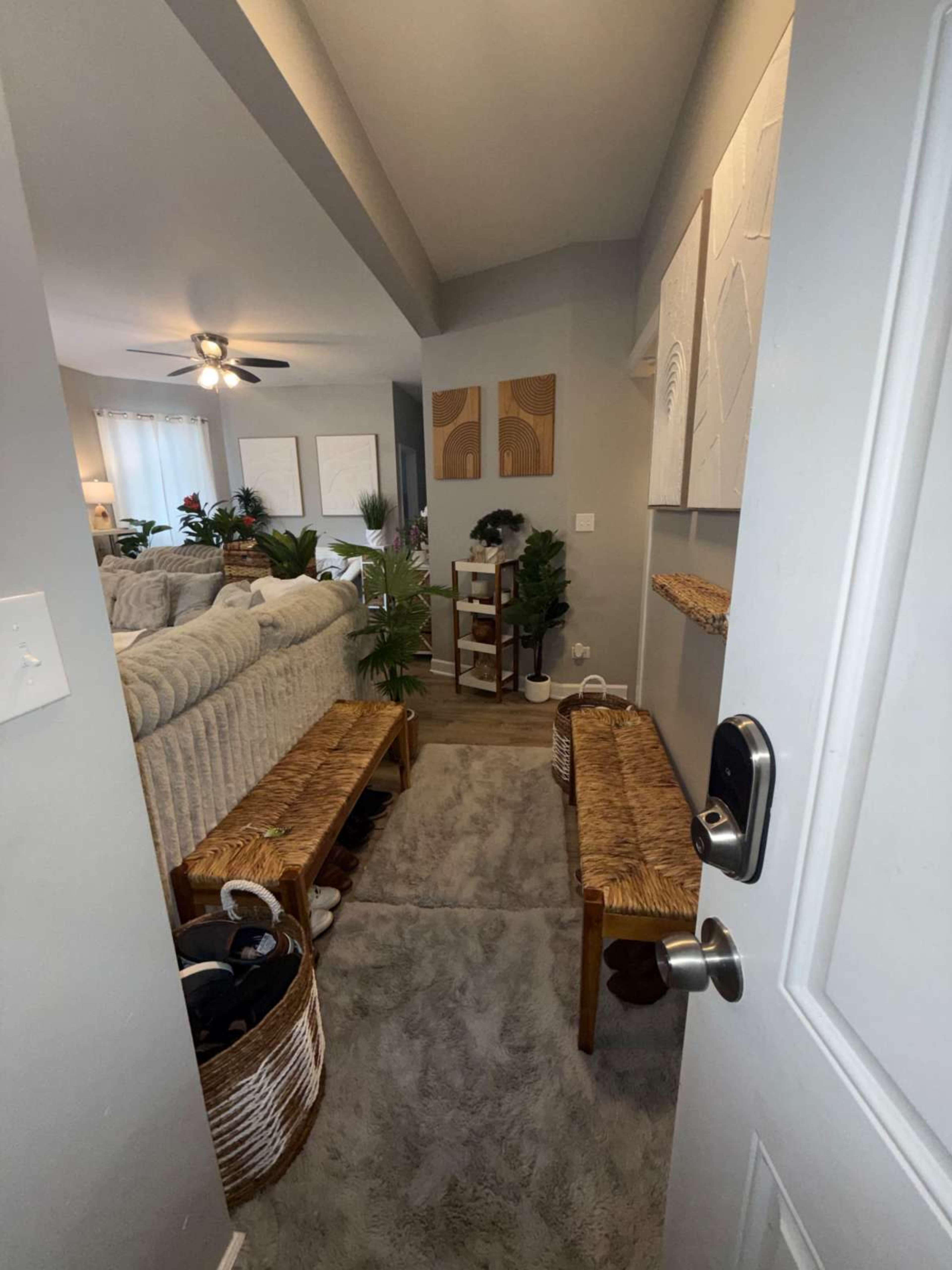 The image shows a hallway entryway with two wooden benches, a small shelf displaying decorative items, and a living area visible in the background, adorned with plants and a cozy sofa.