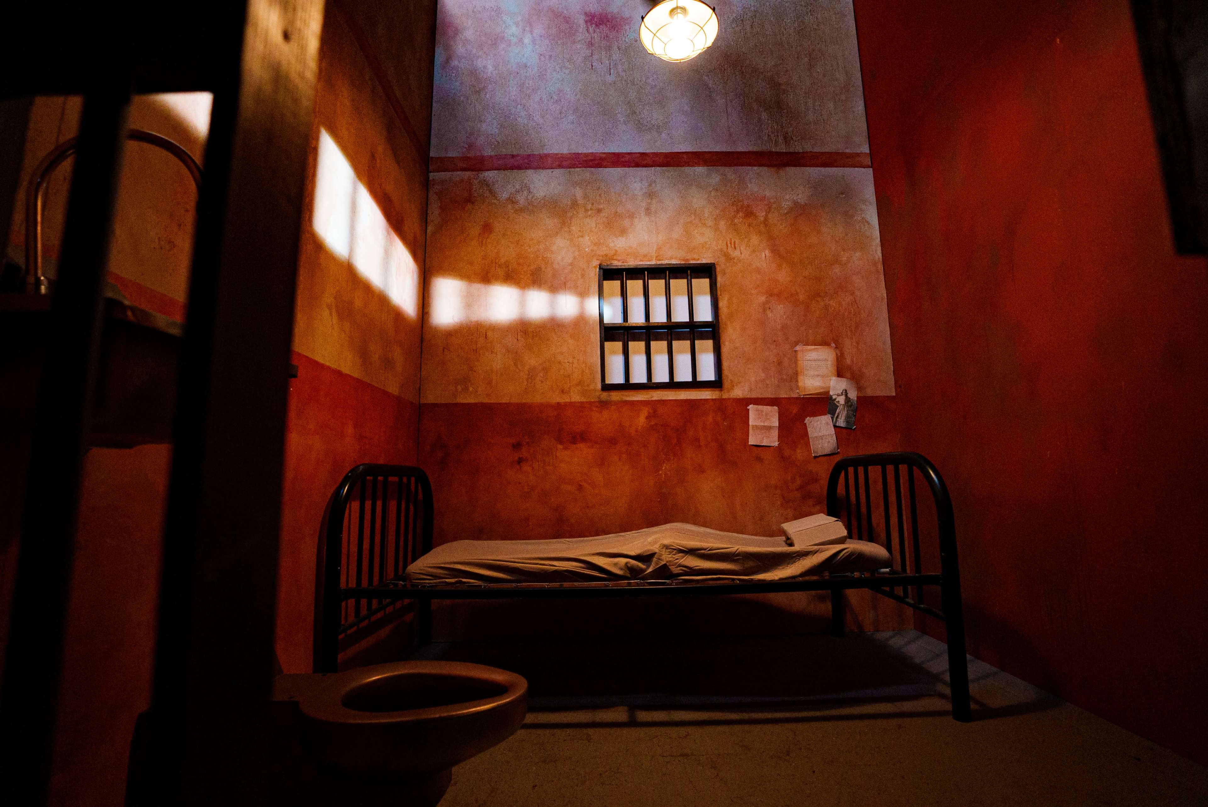 A sparse prison cell featuring a metal bed, a small barred window, and a toilet.