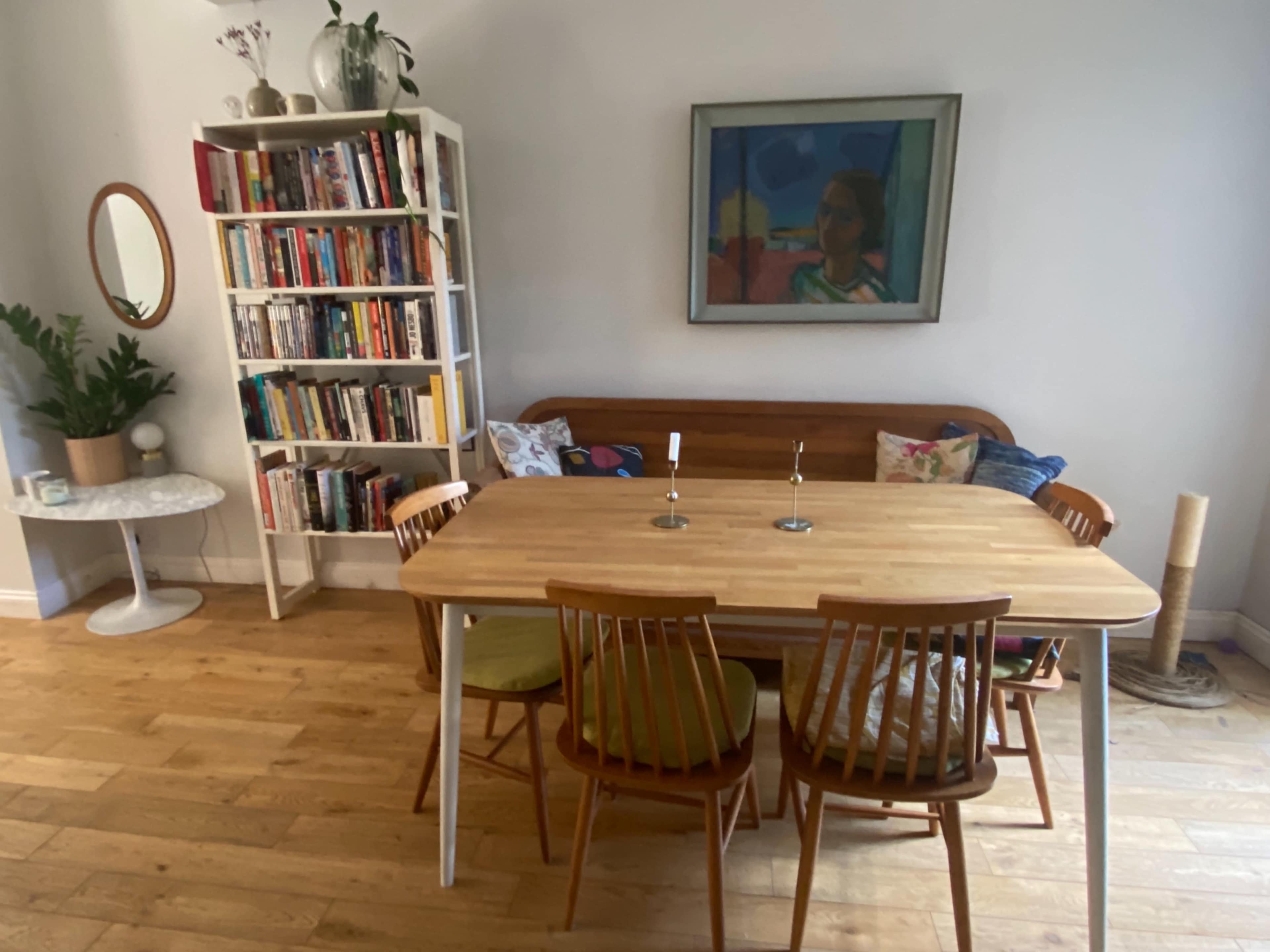 A dining area features a wooden table with four chairs, a bookshelf filled with books, and a colorful painting on the wall.