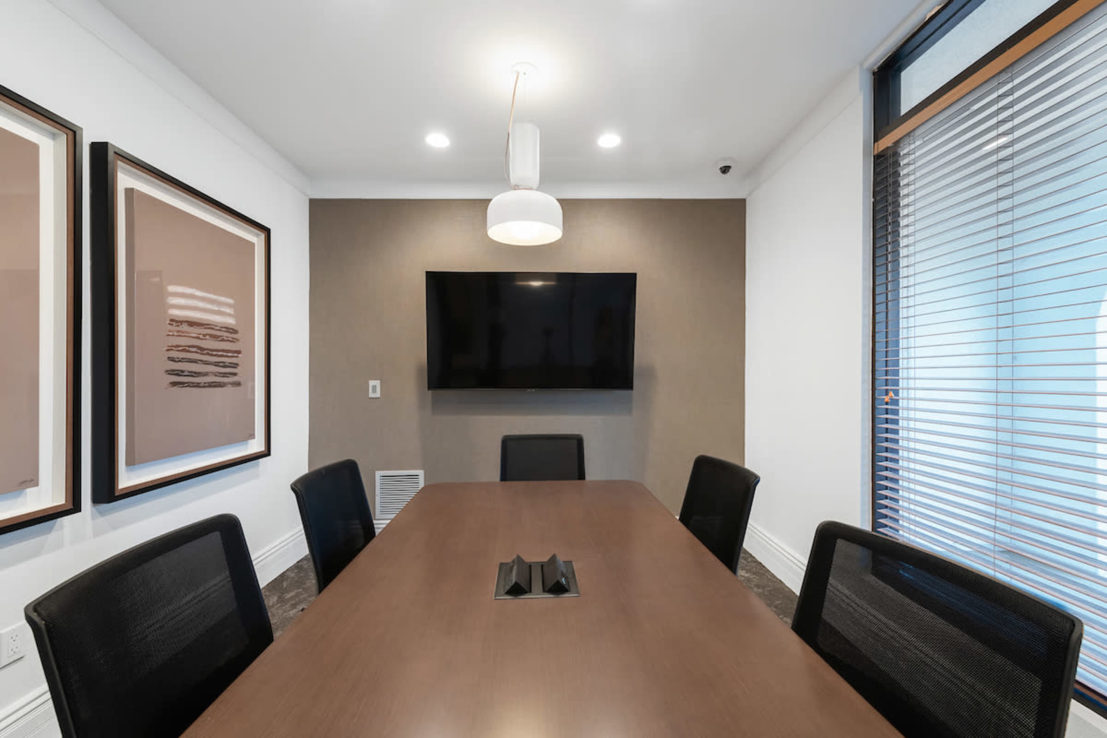 A modern conference room features a wooden table, six black chairs, a wall-mounted television, and framed artwork.
