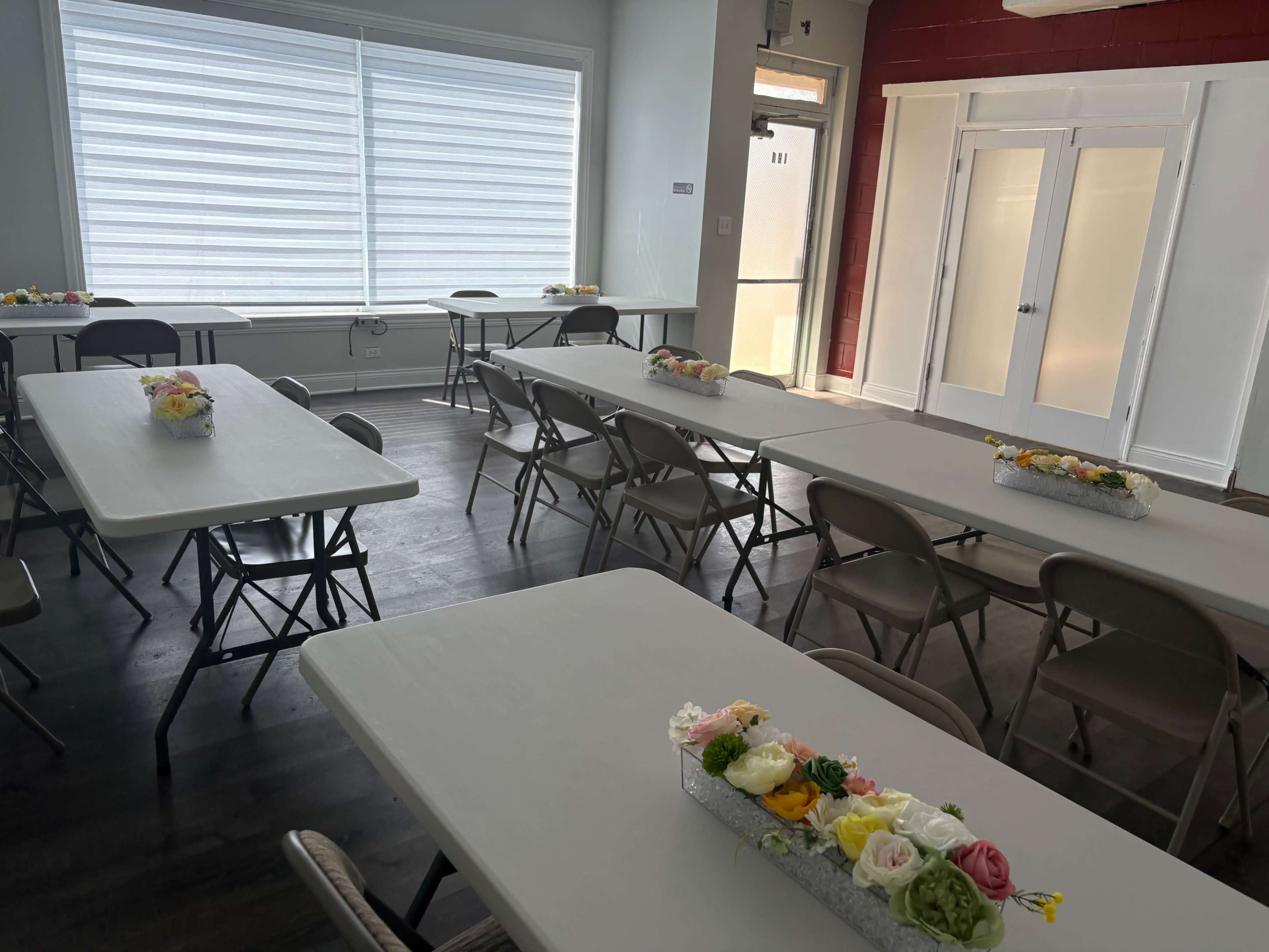The image shows a room set up with several tables and chairs arranged for an event, decorated with floral centerpieces.