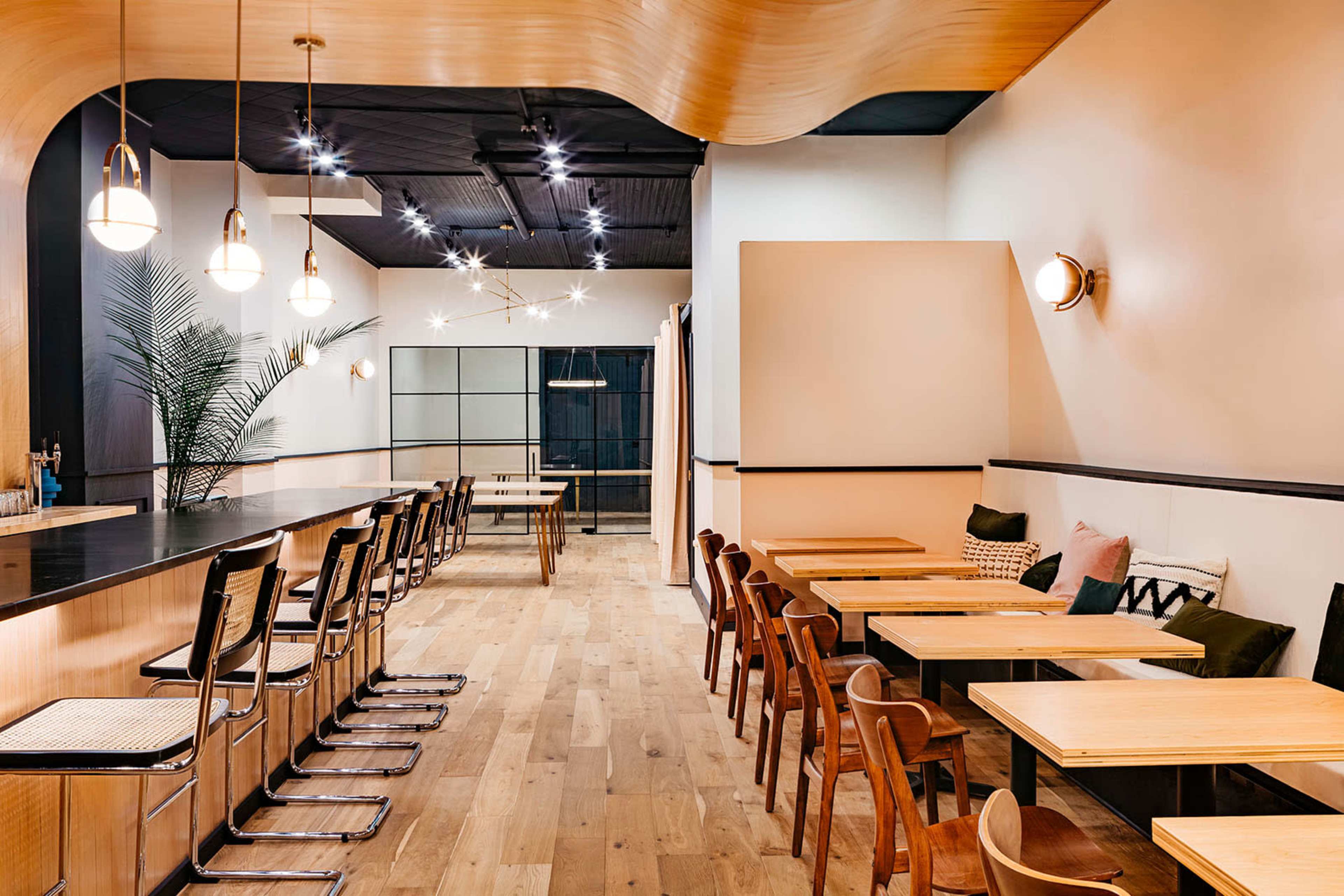 A modern café interior featuring a long bar with stools and a row of wooden tables and chairs, illuminated by pendant lights.