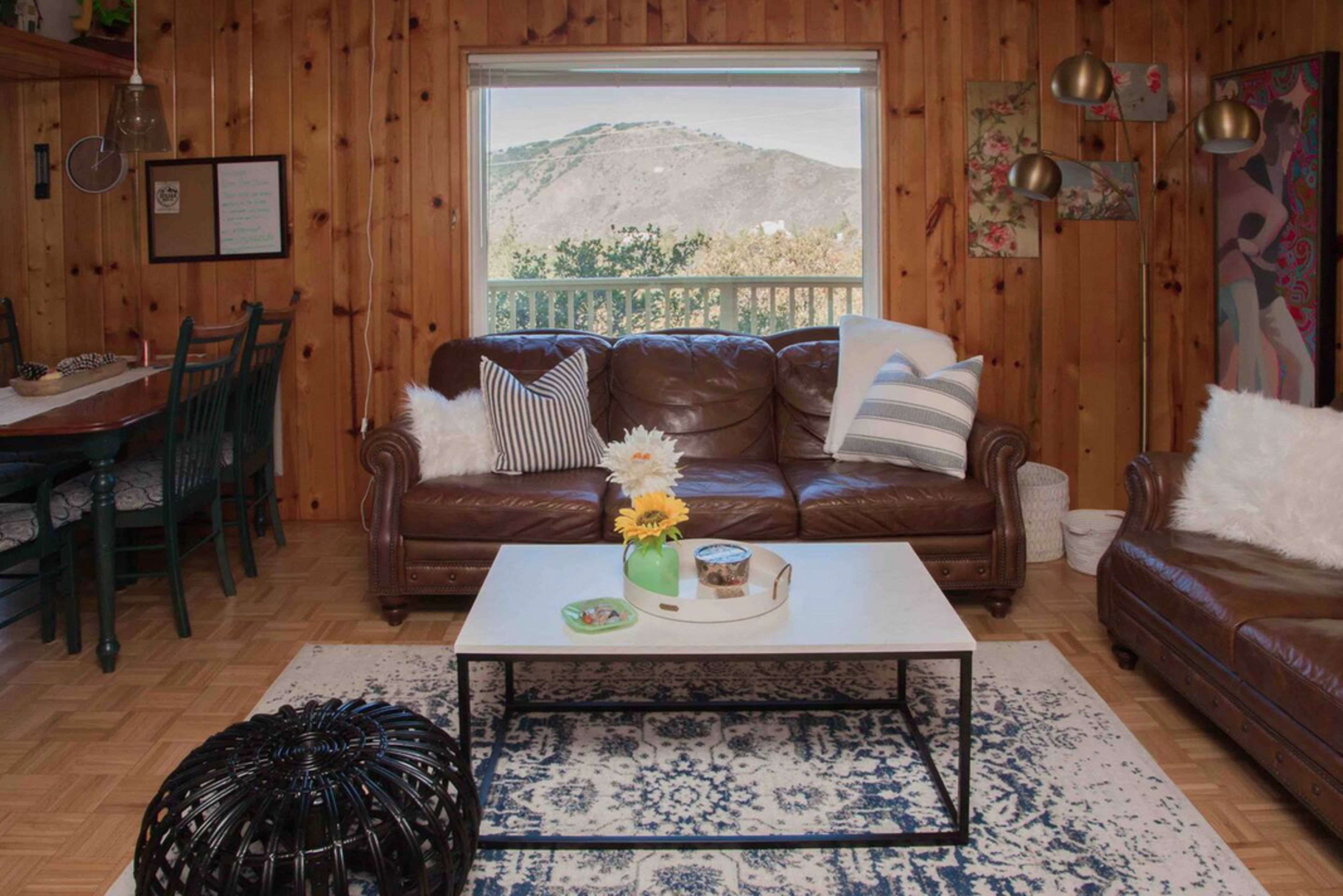 A cozy living room with a brown leather sofa, a coffee table, and a large window displaying a mountain view.