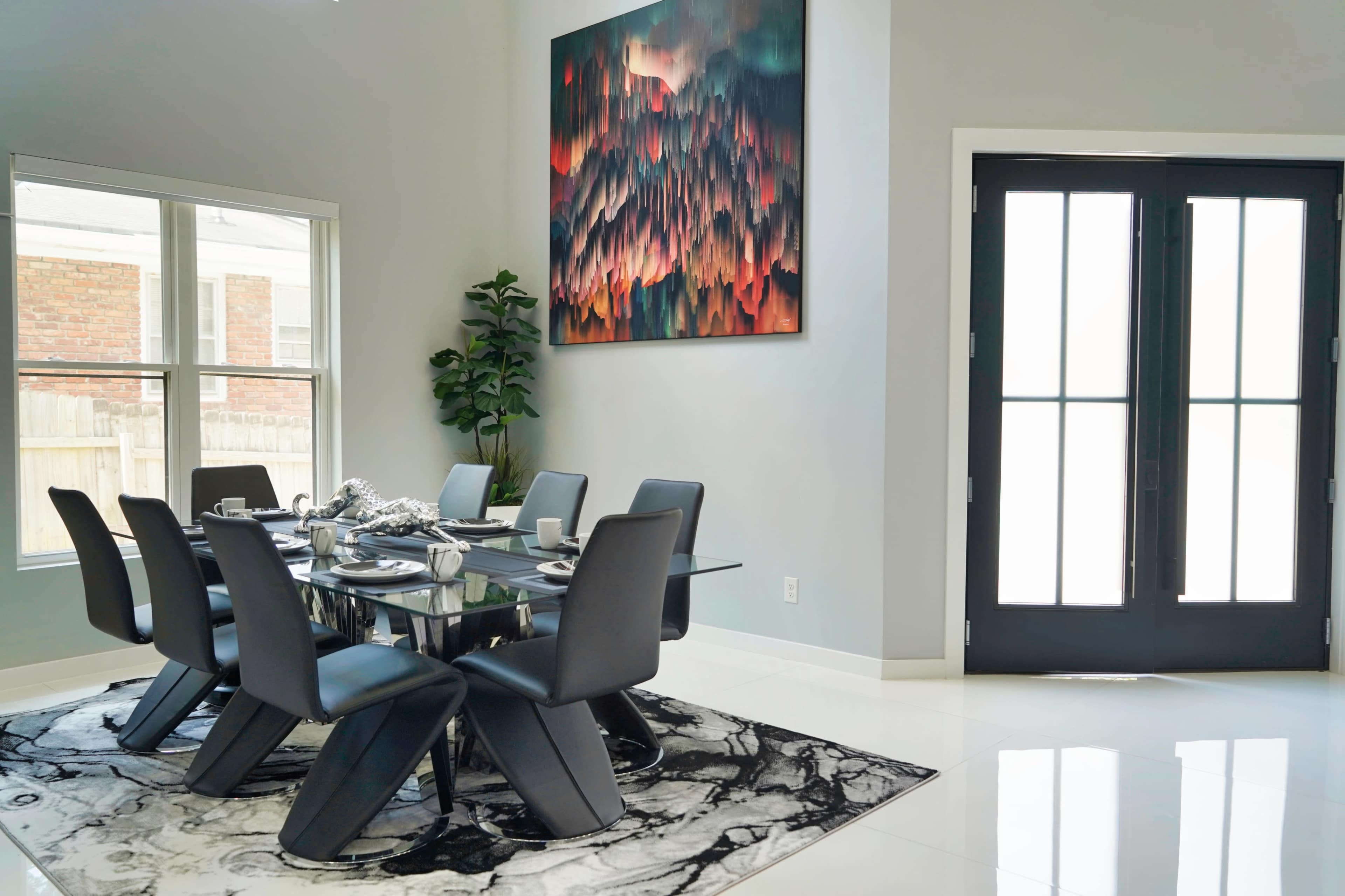 A modern dining area featuring a glass table surrounded by black chairs, with a large artwork on the wall and a plant in the corner near a doorway.