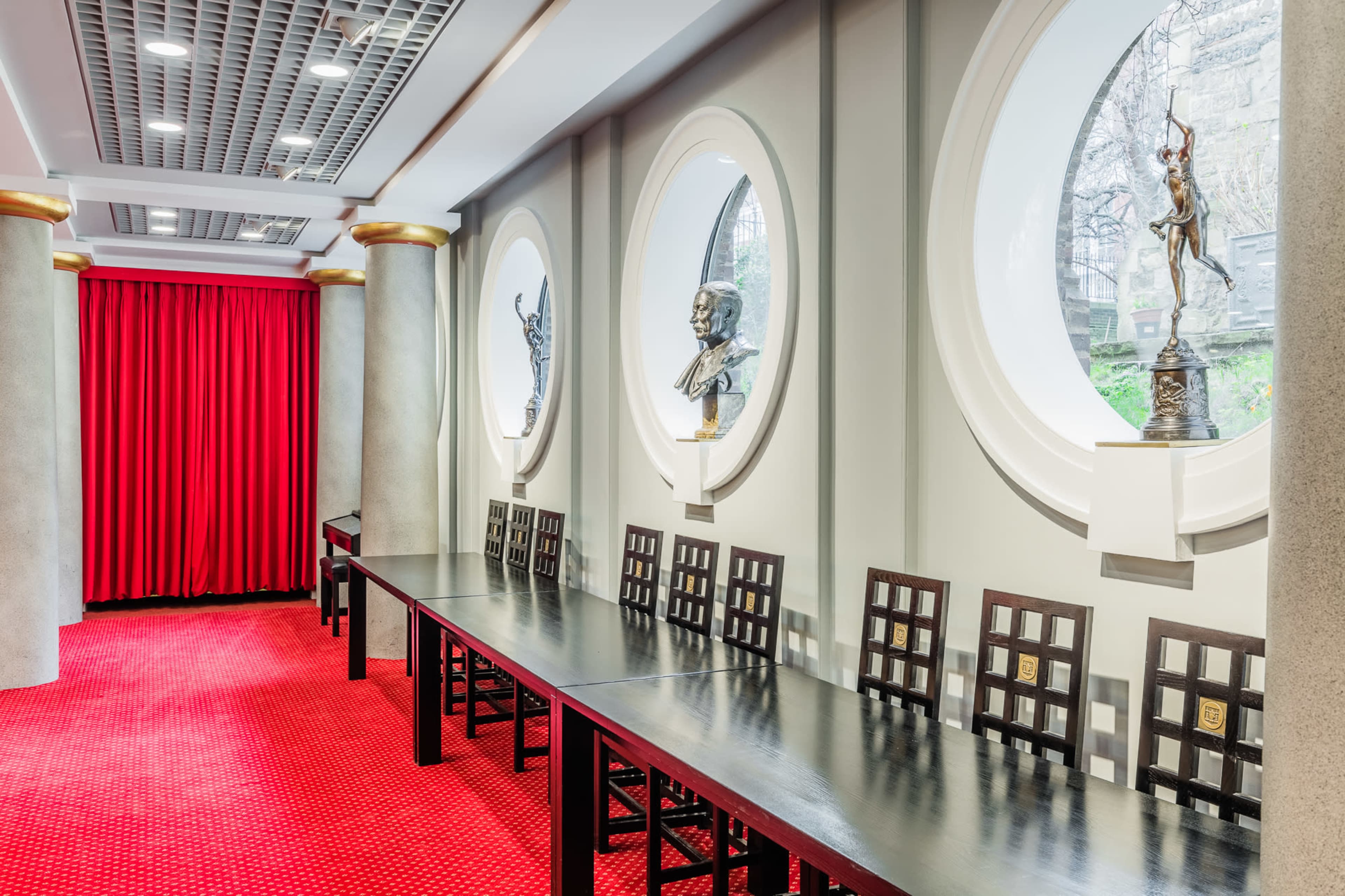 A long, dark table is set against a wall with circular windows and bronze sculptures, surrounded by red curtains and carpet.
