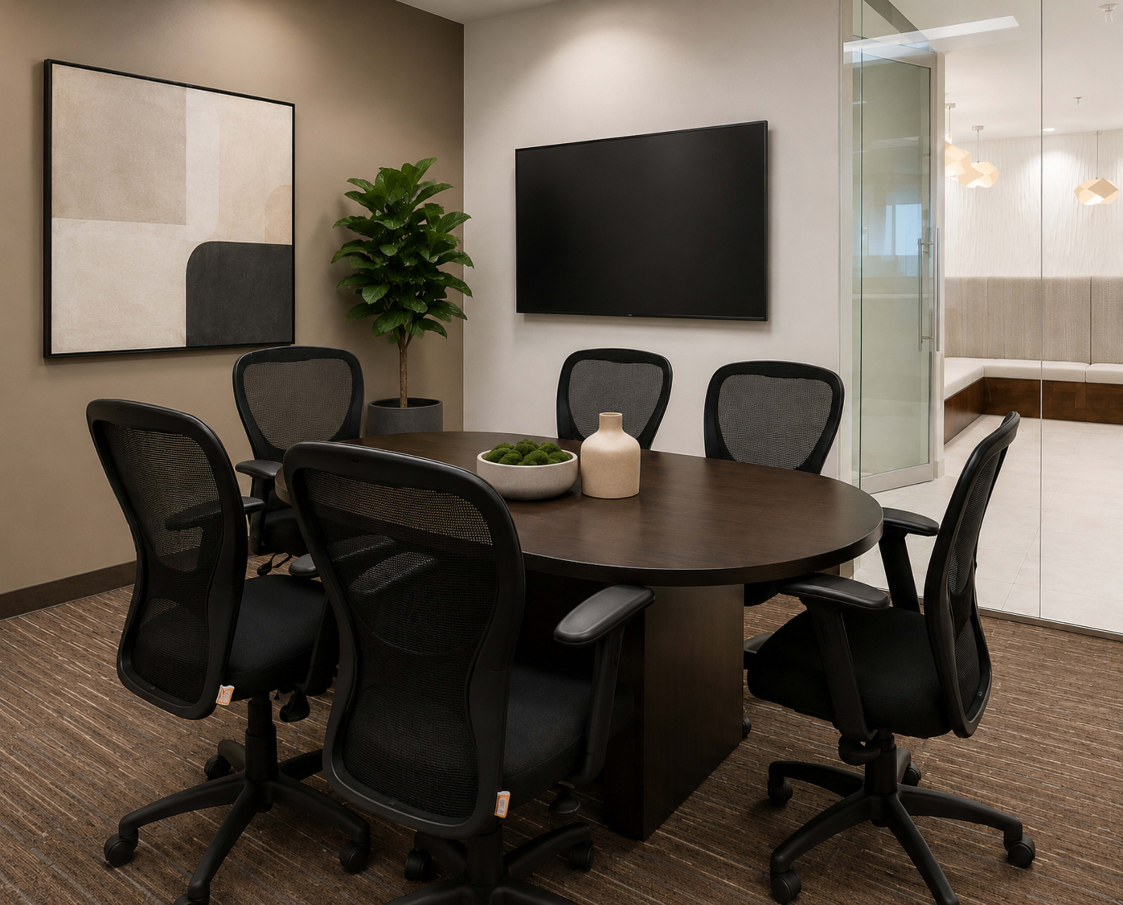 A modern conference room features a round brown table surrounded by six black ergonomic chairs, with a decorative plant, a minimalist wall art piece, and a TV mounted on the wall.