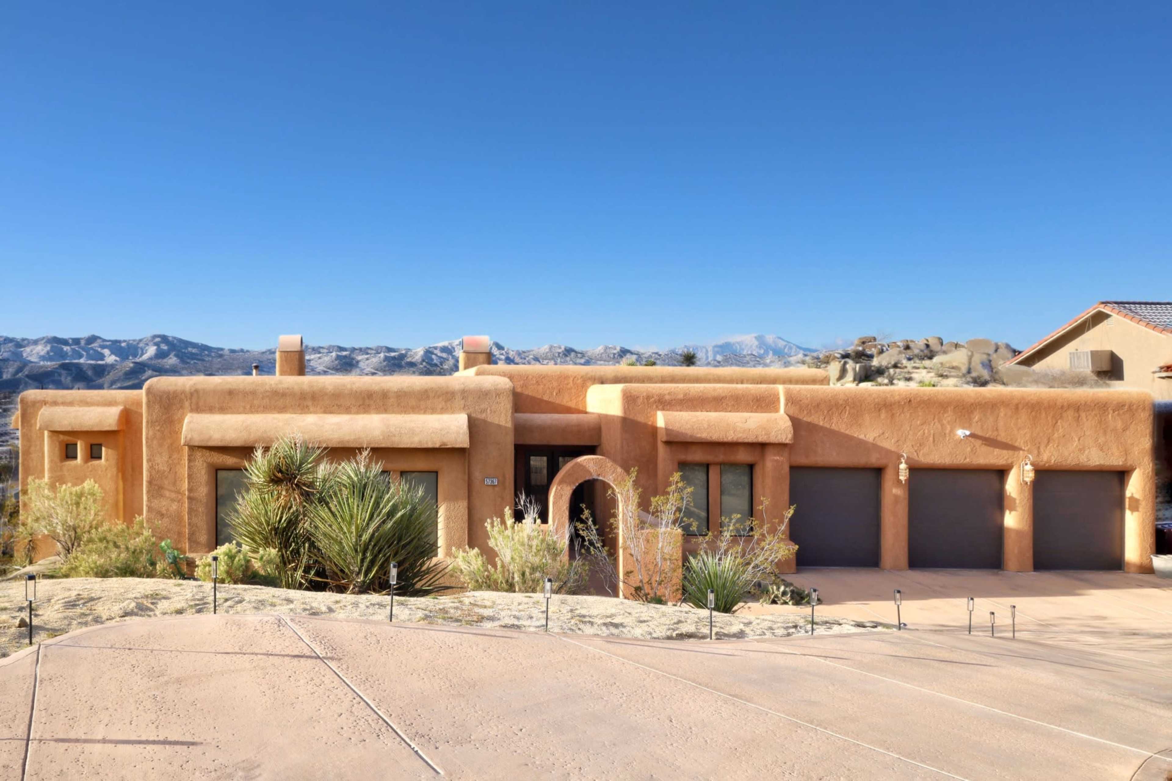 A single-story adobe-style house with three garage doors is situated in a desert landscape, surrounded by low vegetation and mountains in the background.
