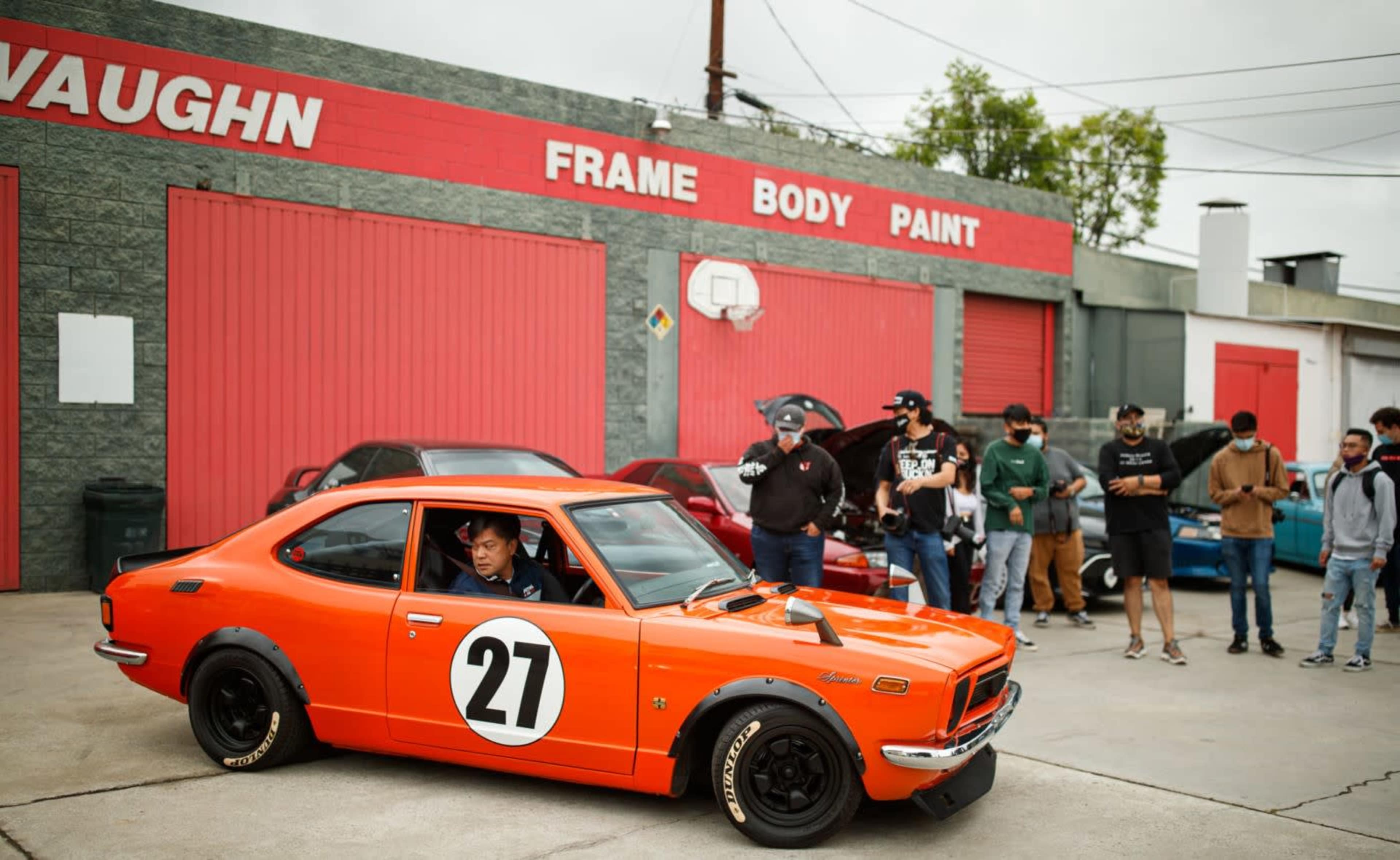 A bright orange car with the number 27 on its side is parked outside a body shop, while a group of people gathers nearby.