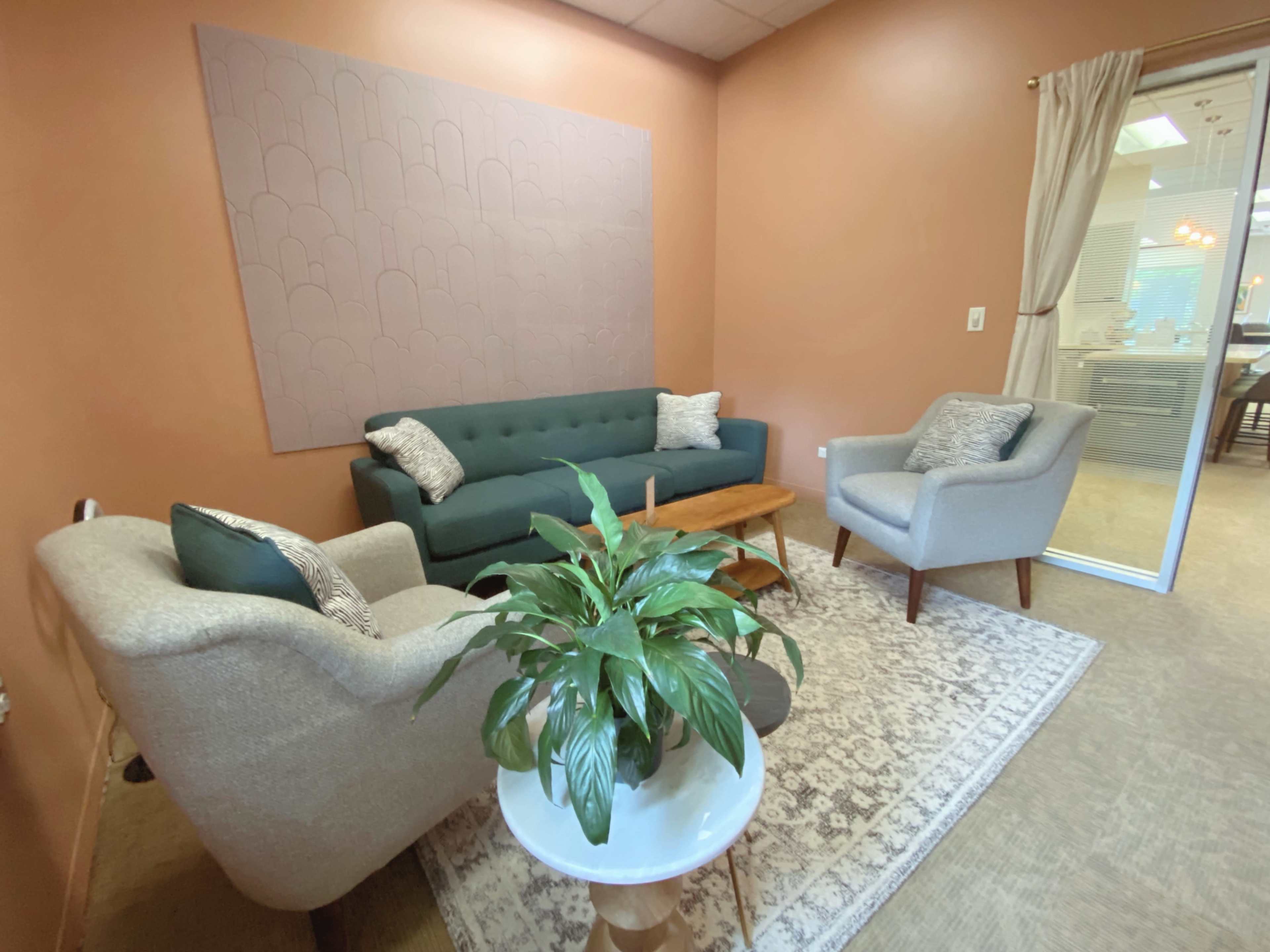 The image shows a small room with two upholstered armchairs, a green sofa, a wooden coffee table, and a large plant on a round table, all set against a peach-colored wall.