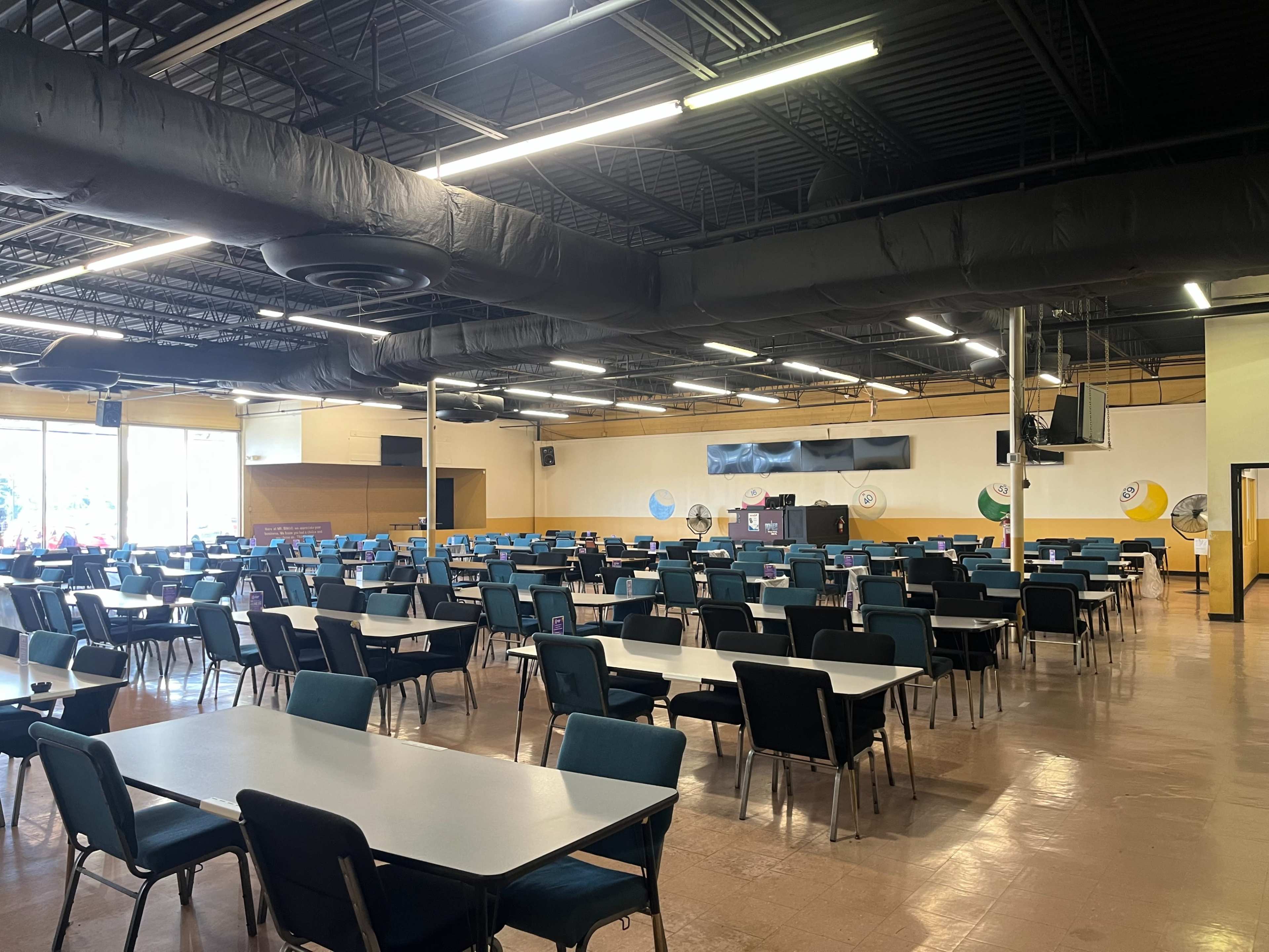 A spacious dining hall filled with rows of tables and chairs, with large windows providing natural light.