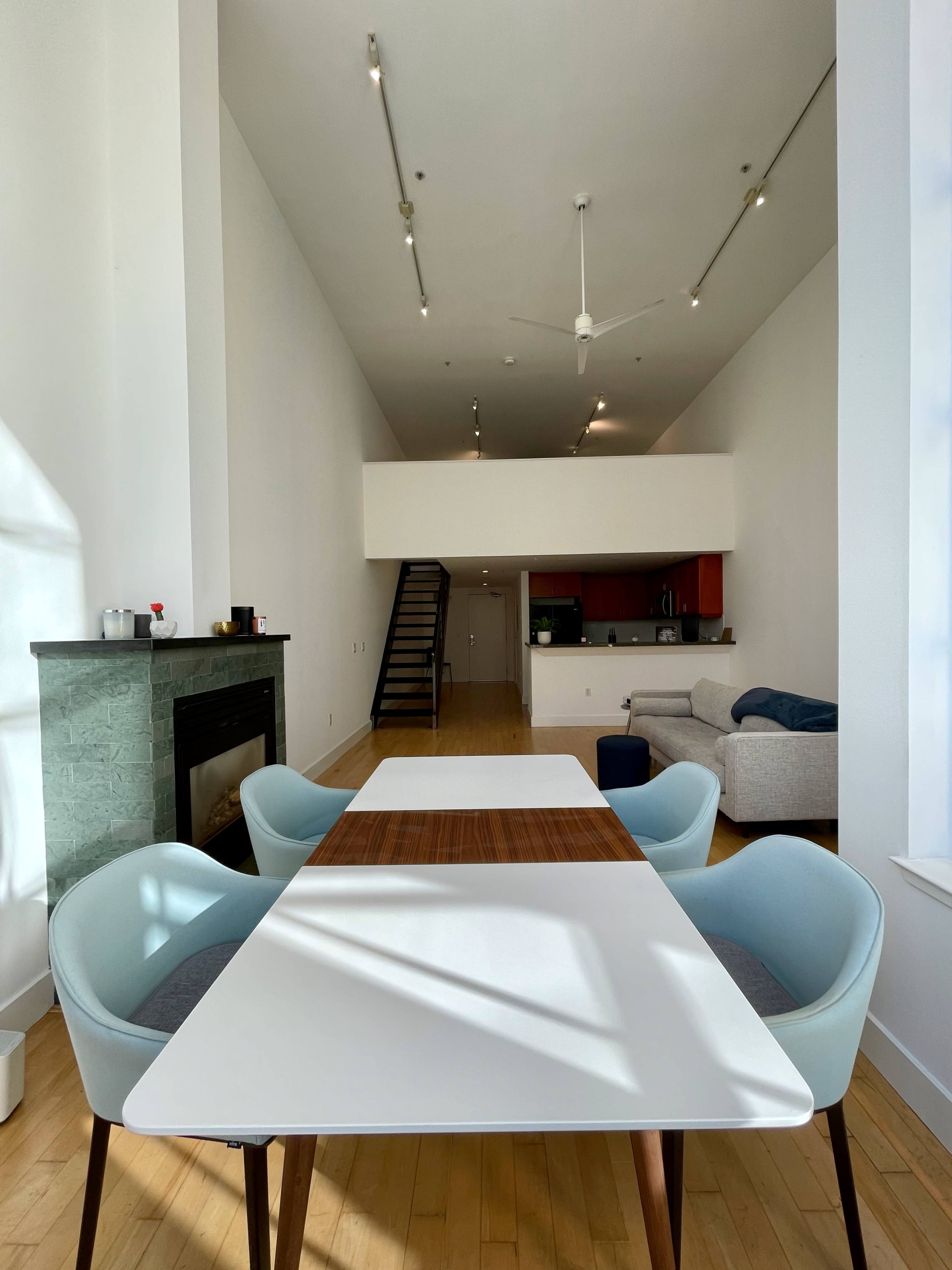 A spacious modern room features a white table with light blue chairs, a green fireplace, and a staircase leading to an upper level.