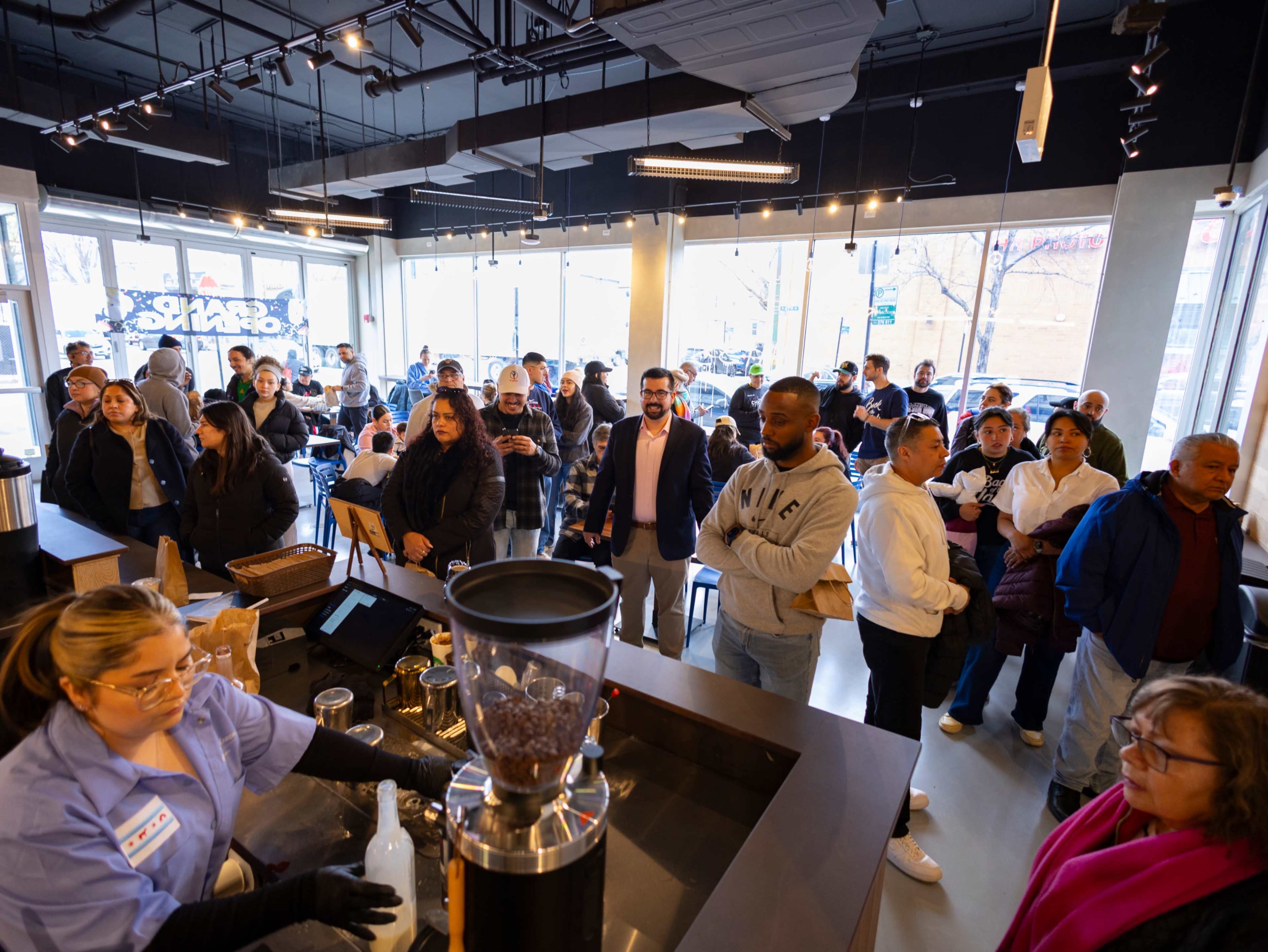 A large crowd of people gathers inside a coffee shop, with a barista preparing drinks at the counter.