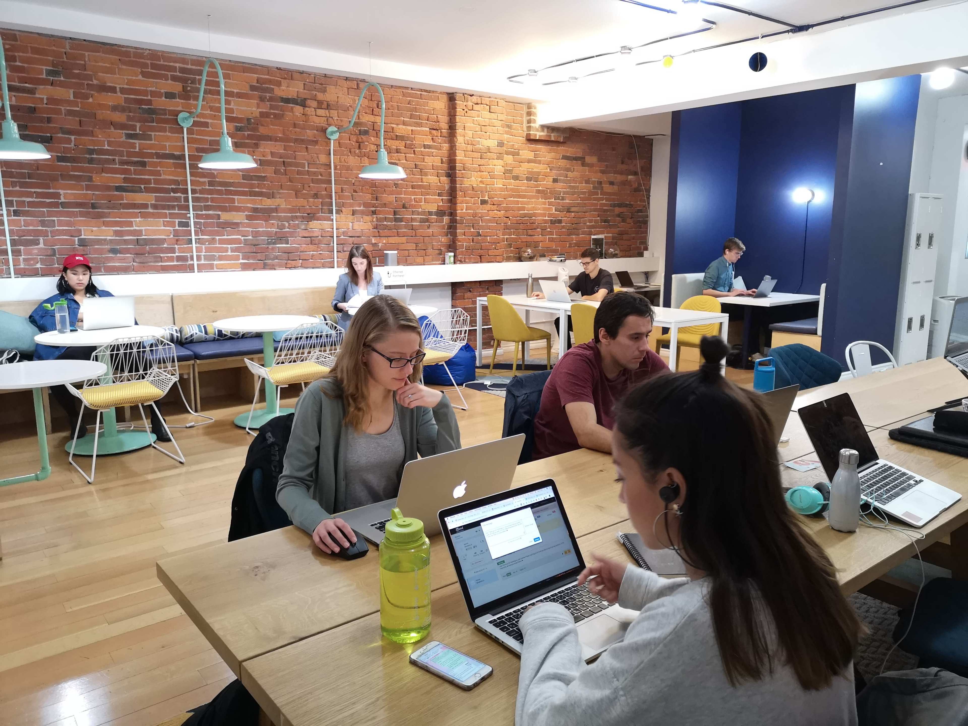 A group of people work on laptops in a modern, open workspace featuring wooden tables and exposed brick walls.