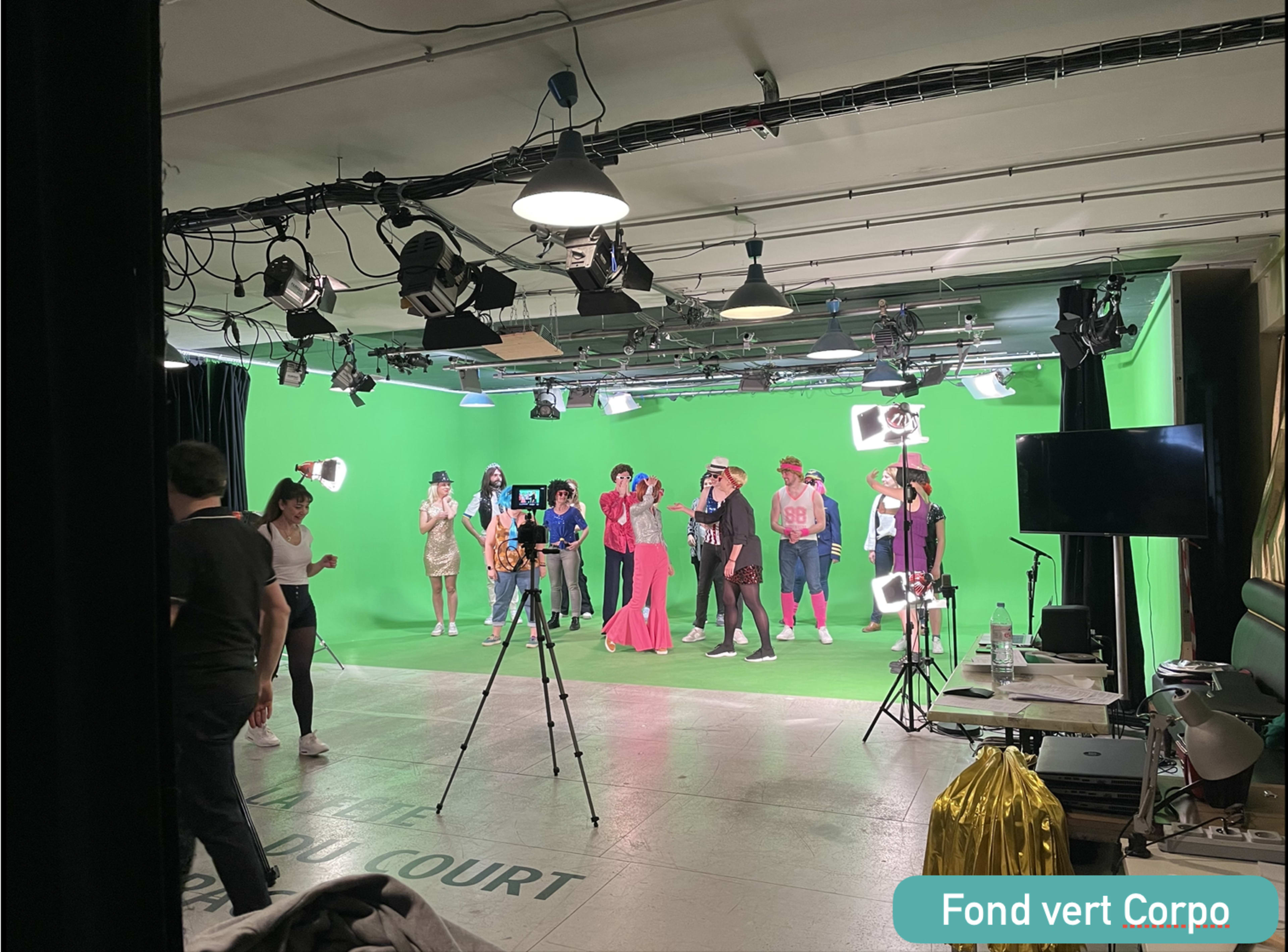 A group of people dressed in various costumes stands in front of a green screen in a studio, with cameras and lights set up around them.