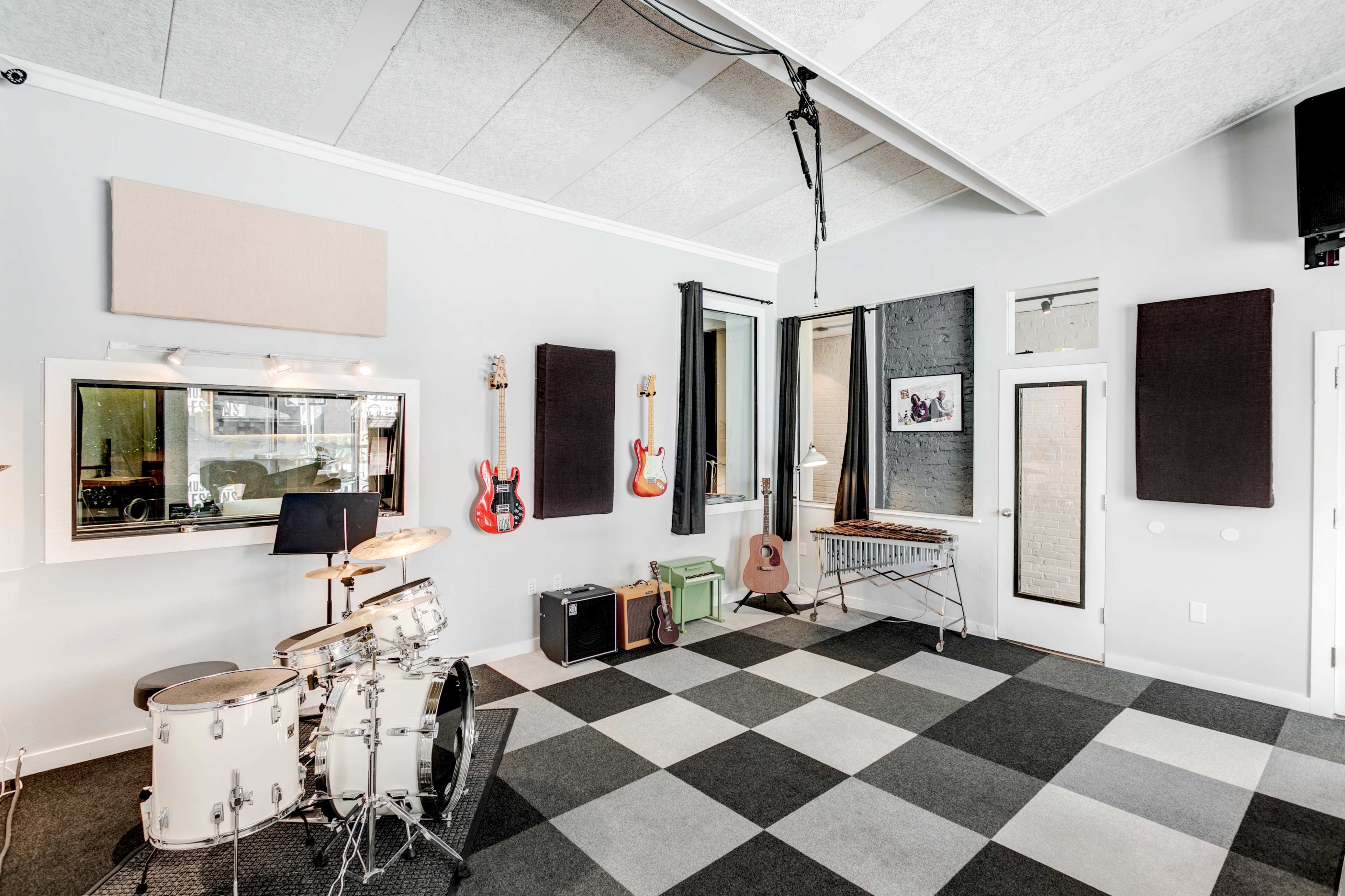 A spacious music studio features a drum set, electric guitars on the wall, and a vibraphone on a stand, with black and white checkered flooring and soundproofing materials.
