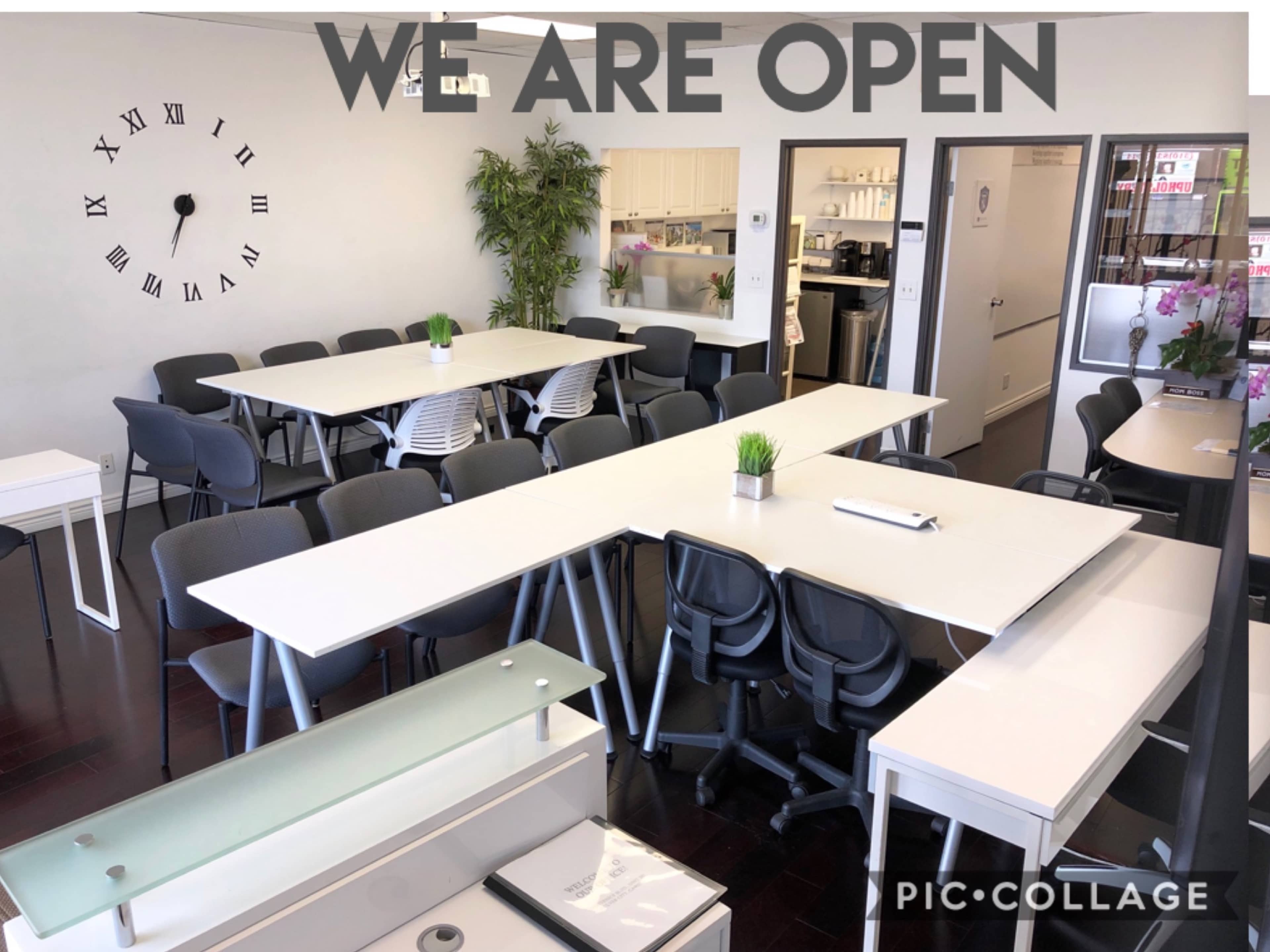 A bright, modern office space with several white tables and black chairs arranged for meetings, featuring a large wall clock and a plant in the corner.
