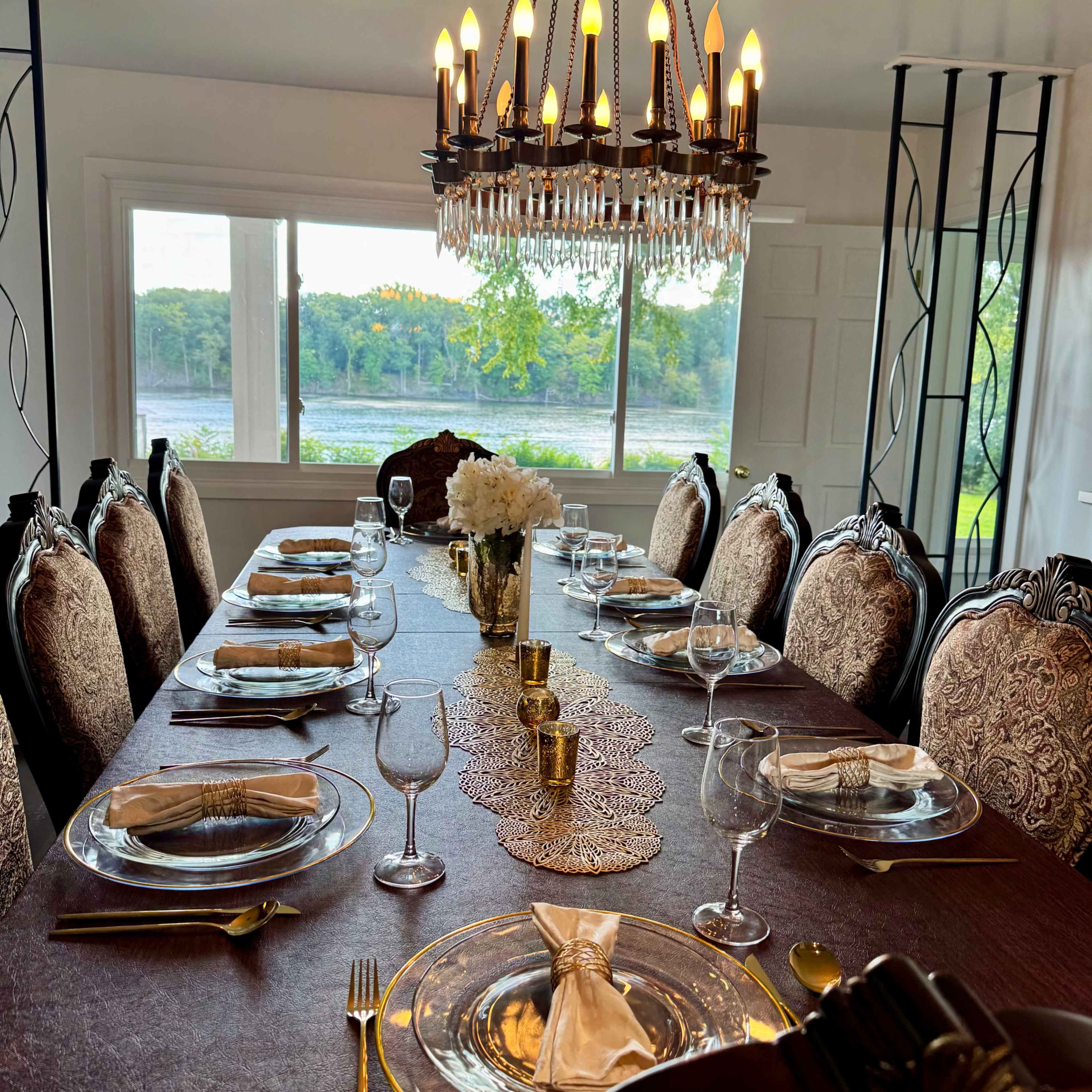 A long dining table is set with elegant plates, glassware, and silverware, surrounded by ornate chairs and illuminated by a chandelier, with a view of a serene landscape outside the window.
