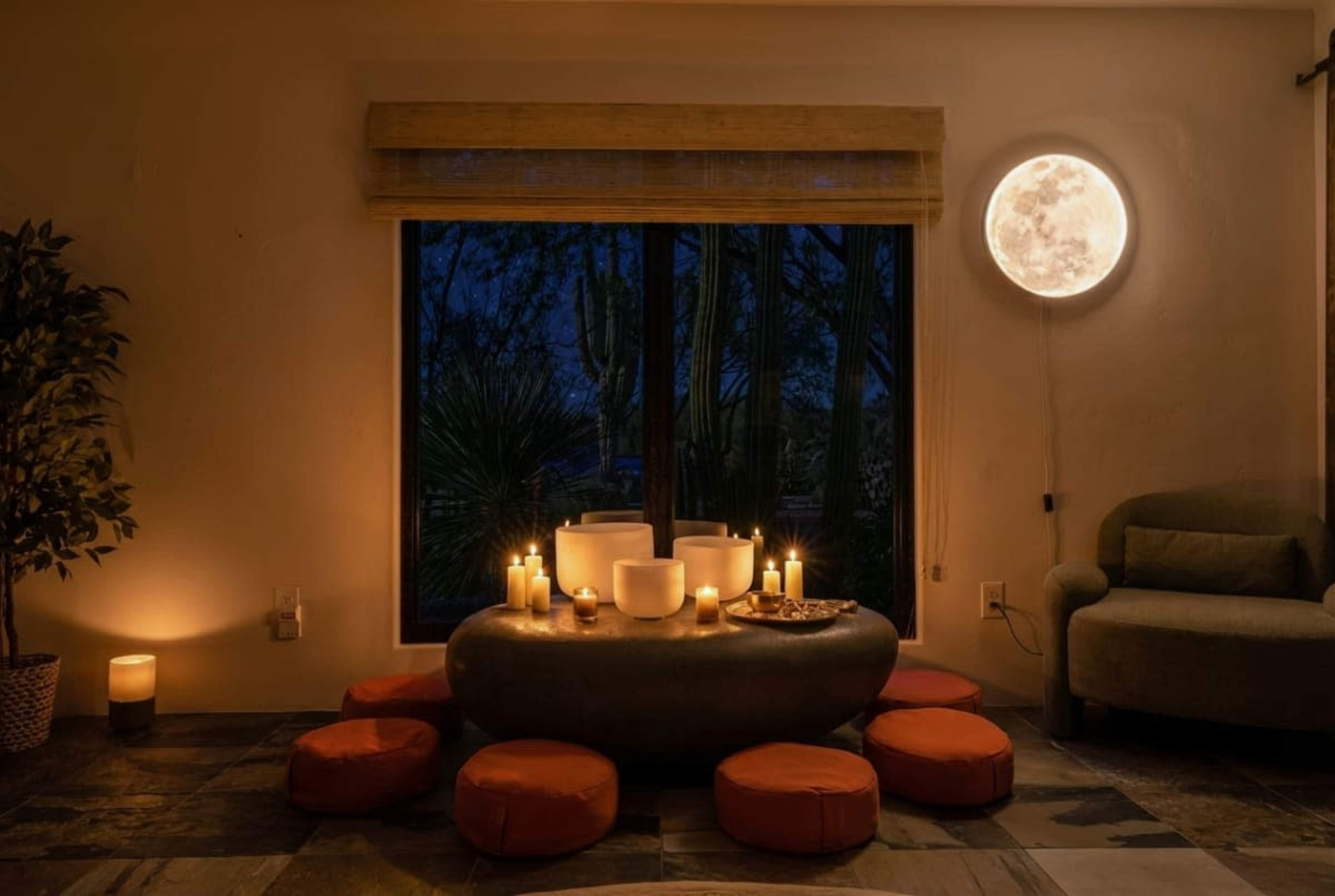 A cozy room features a circular stone table surrounded by orange cushions, with candles and a large moon-shaped light illuminating the space against a darkening backdrop.