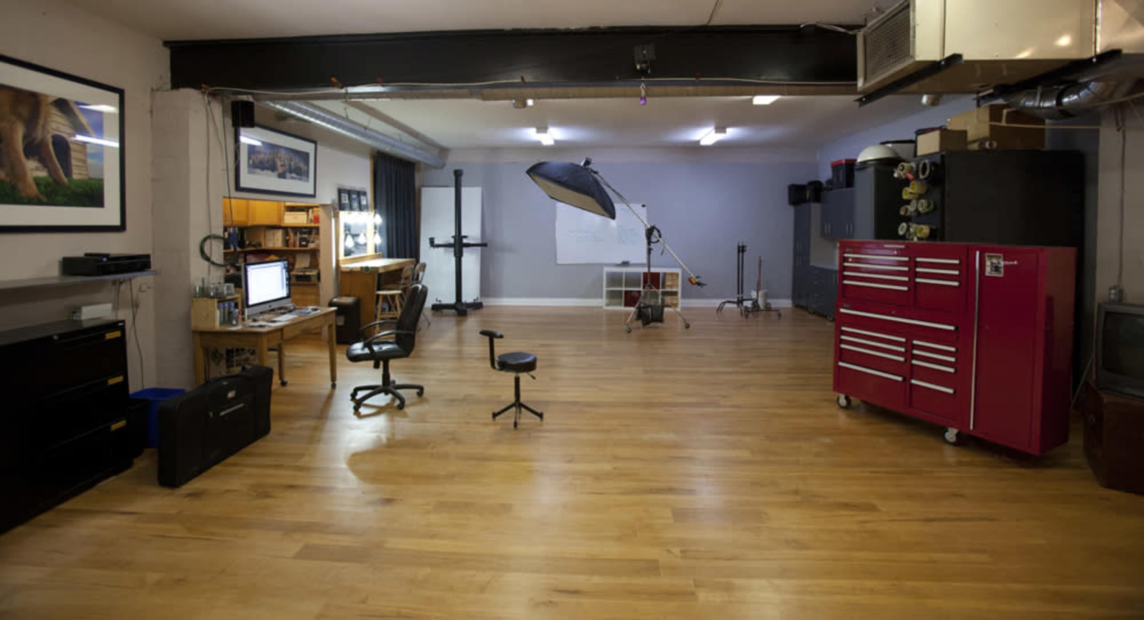 A spacious studio with wooden flooring, featuring a desk with a computer, a rolling chair, and a large red tool chest against the wall, along with various photography equipment set up in the background.