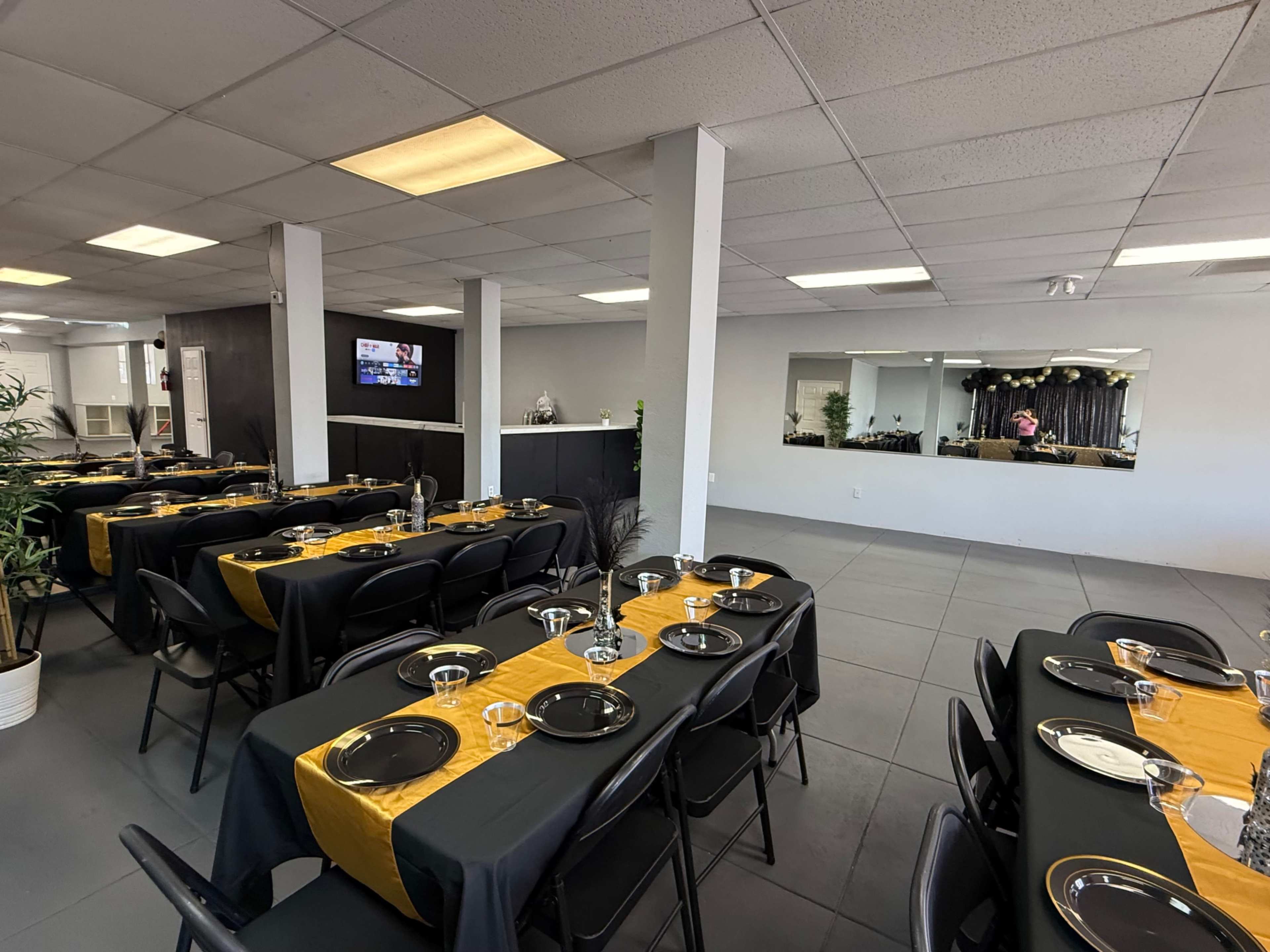 A spacious dining area is set up with several tables covered in black cloth, each laid with plates, cutlery, and glasses, and features a mirrored wall and a television in the background.
