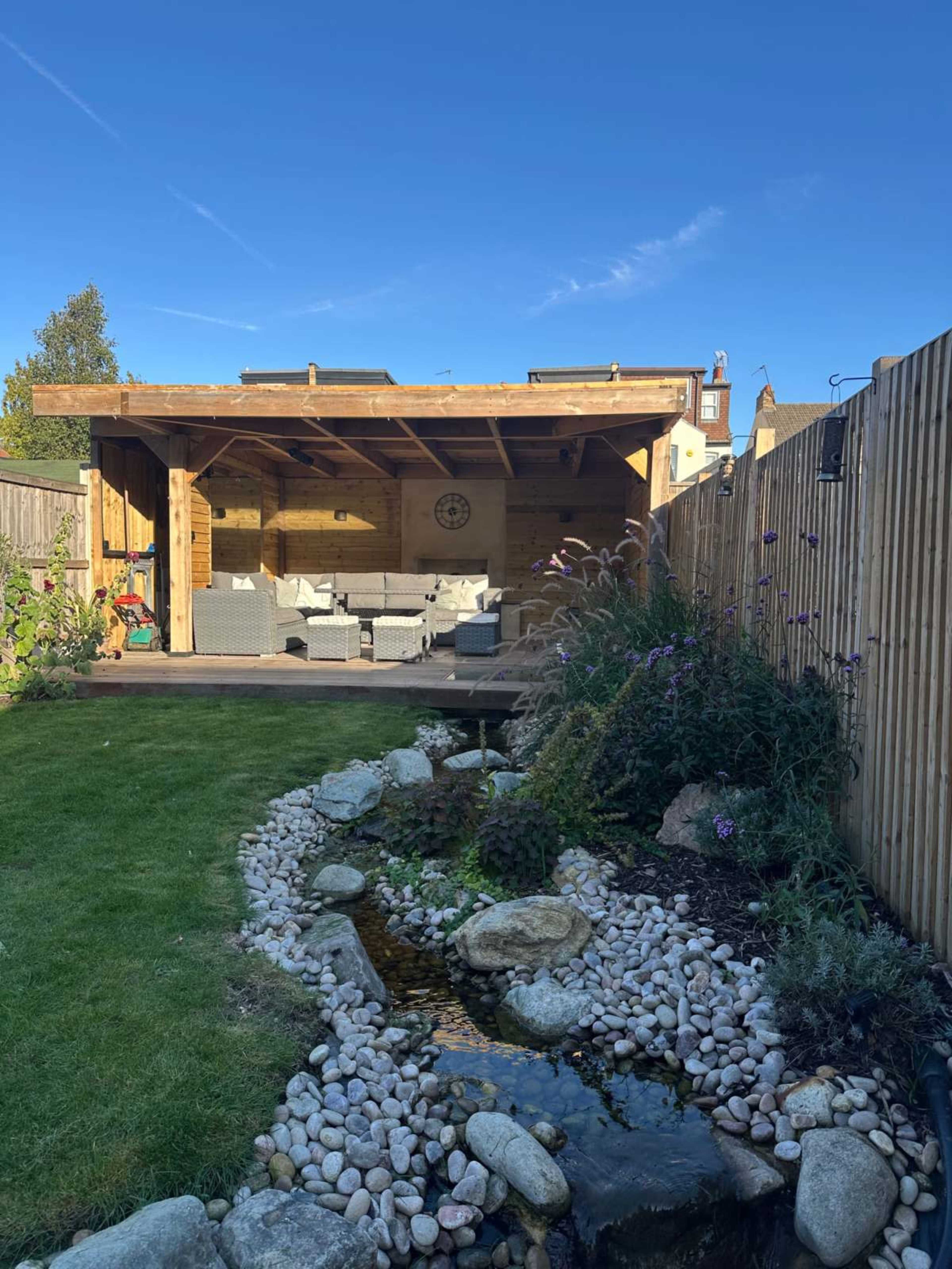 A wooden gazebo with seating is sheltered by a roof, beside a stone-lined garden stream and landscaped plants.