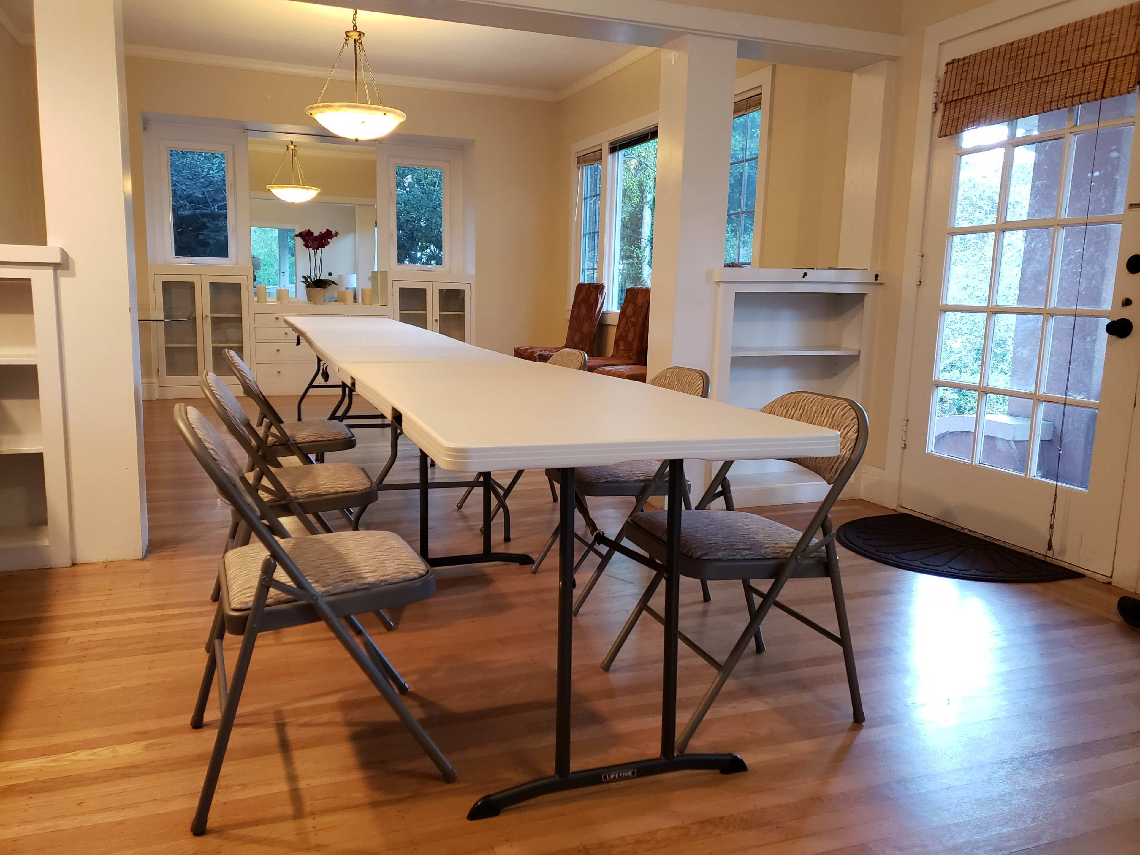 A long white folding table is set up in a bright room with wooden flooring, surrounded by folding chairs and large windows.