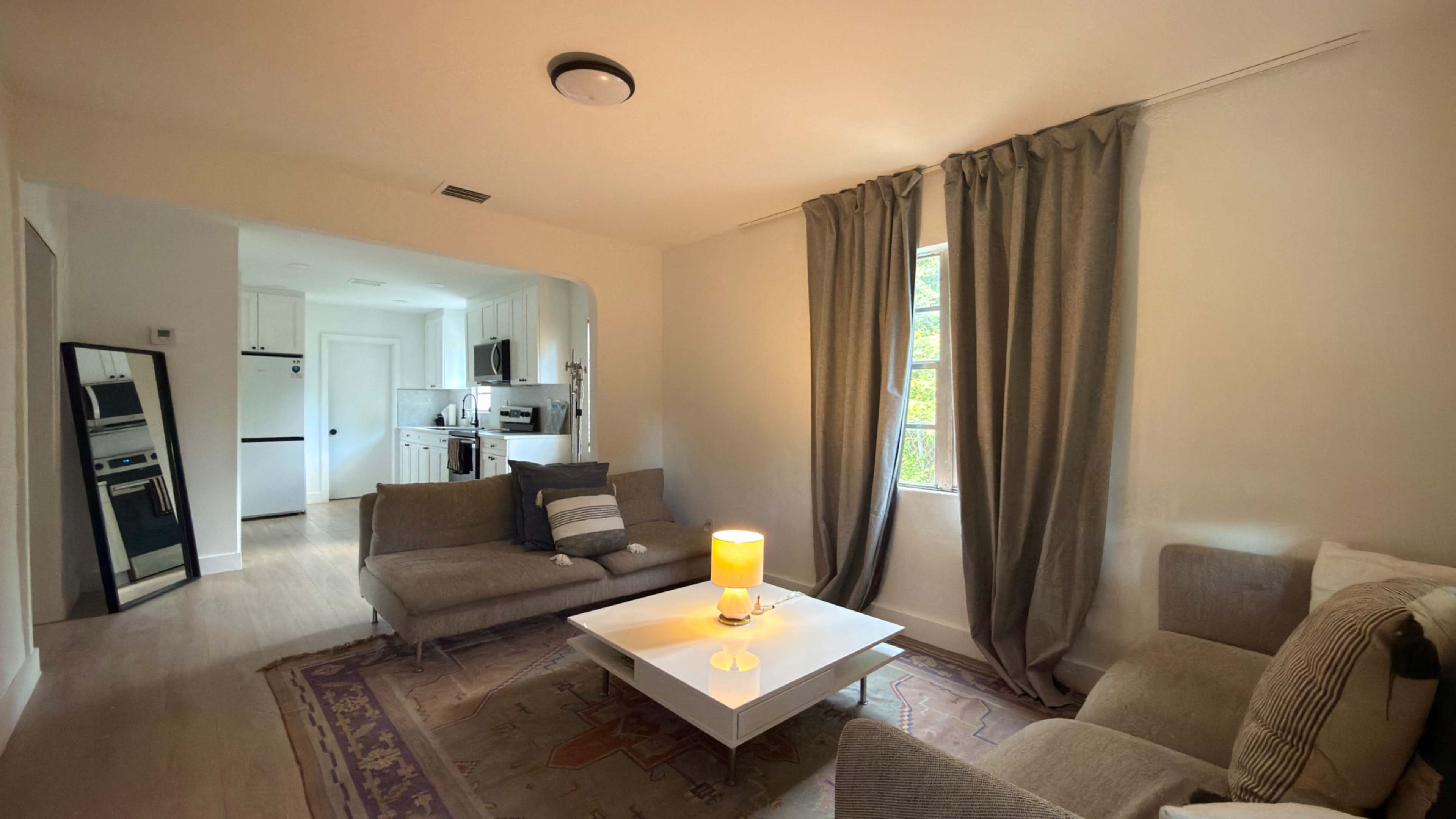 The image shows a living room with two sofas, a coffee table, and a lamp, leading into a kitchenette area in the background.