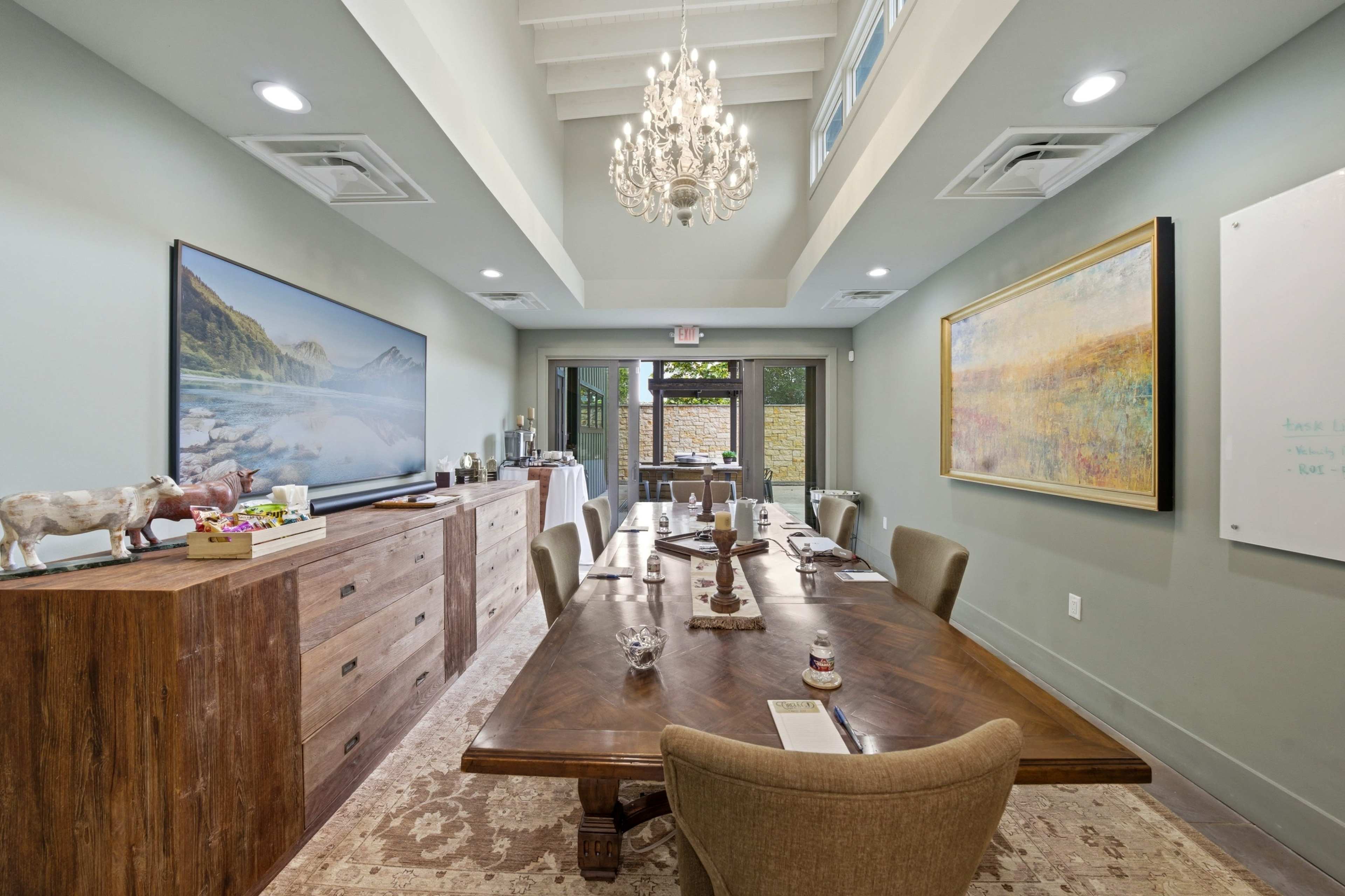 A modern conference room features a wooden table set for a meeting, with a chandelier overhead and artwork on the walls.