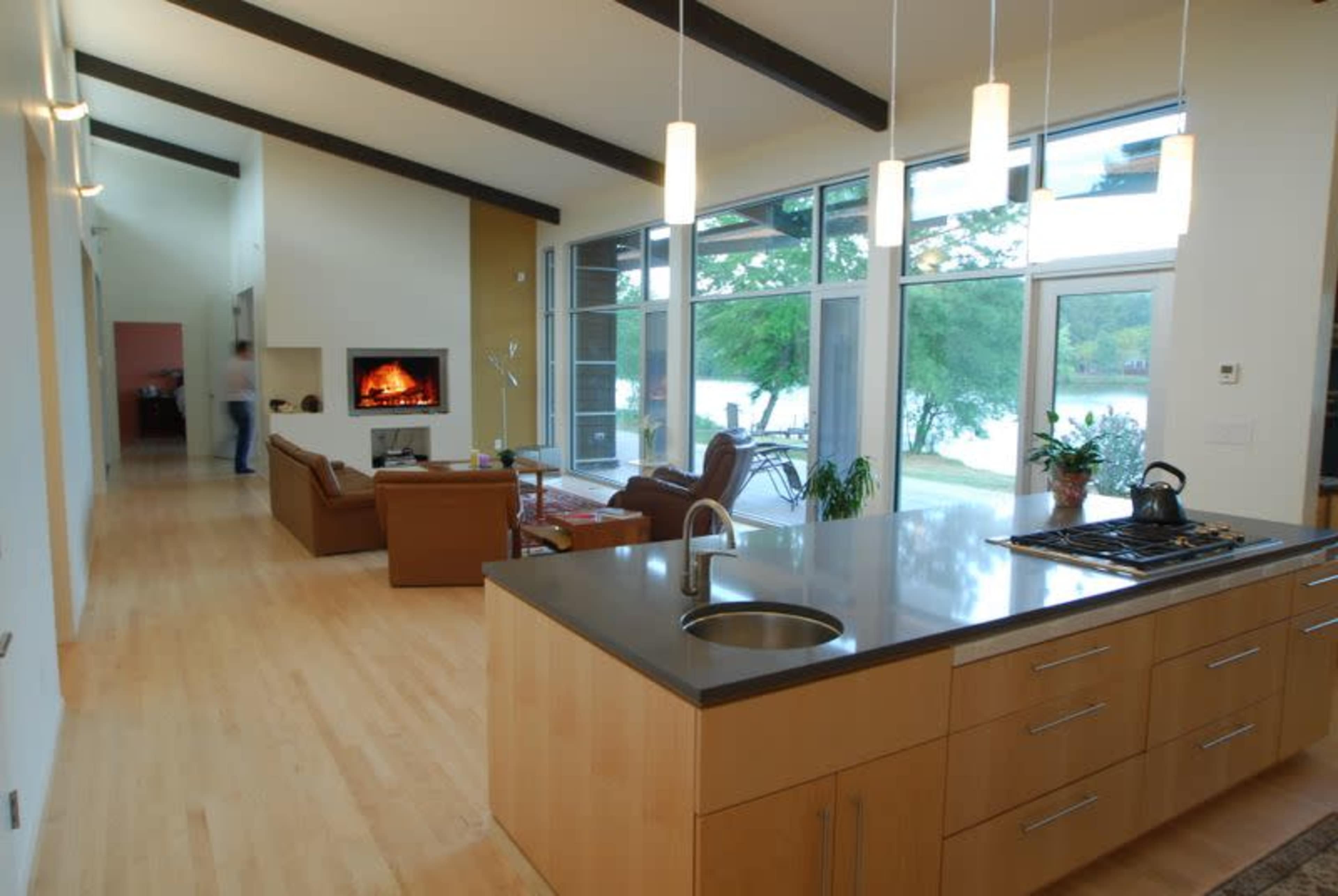 The image shows a modern open-concept kitchen and living area with large windows overlooking a lake, featuring a central island and a fireplace in the living space.