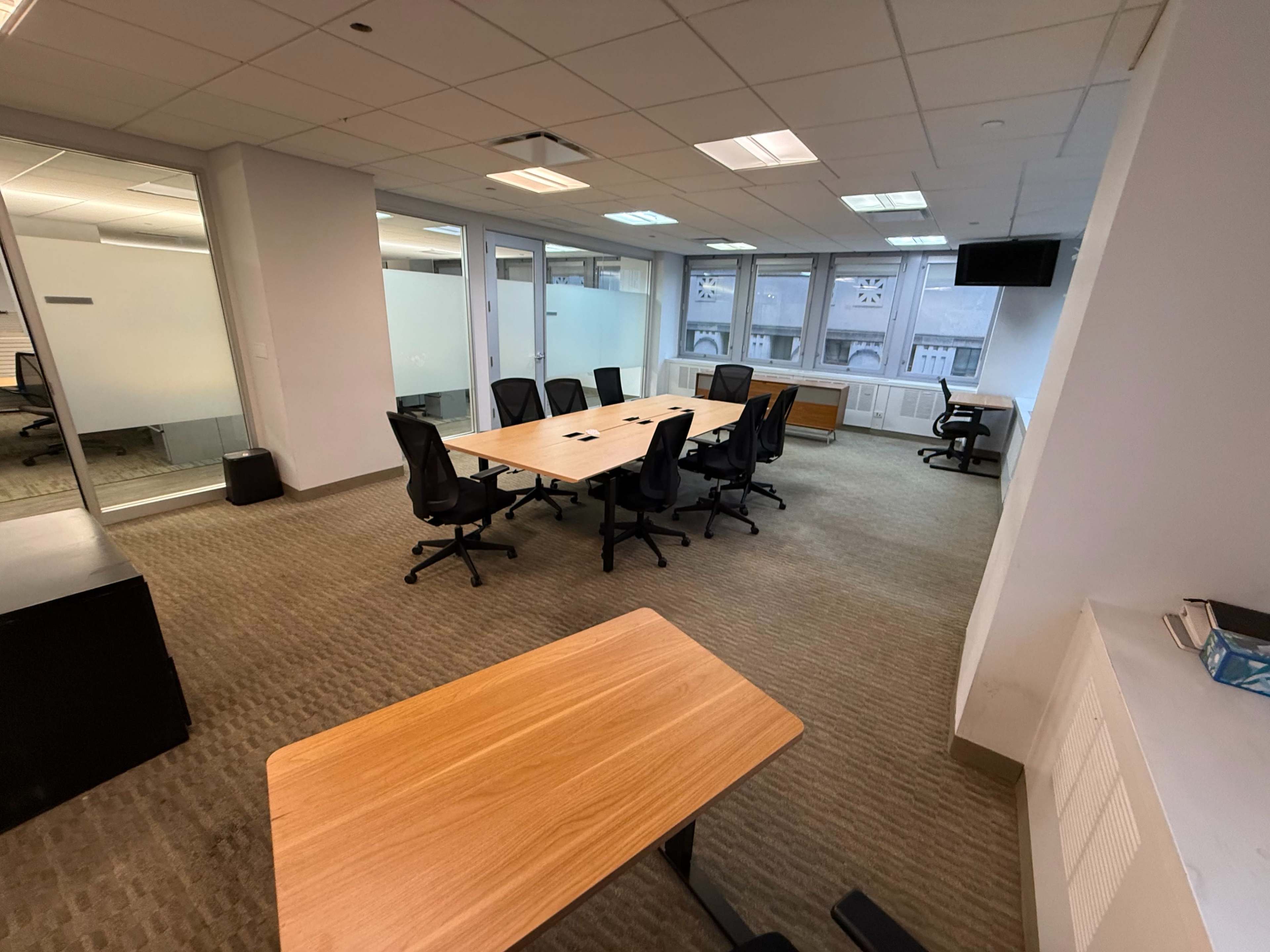 The image shows a large, empty office meeting room with a central wooden table surrounded by several black chairs, and glass partitioned walls.