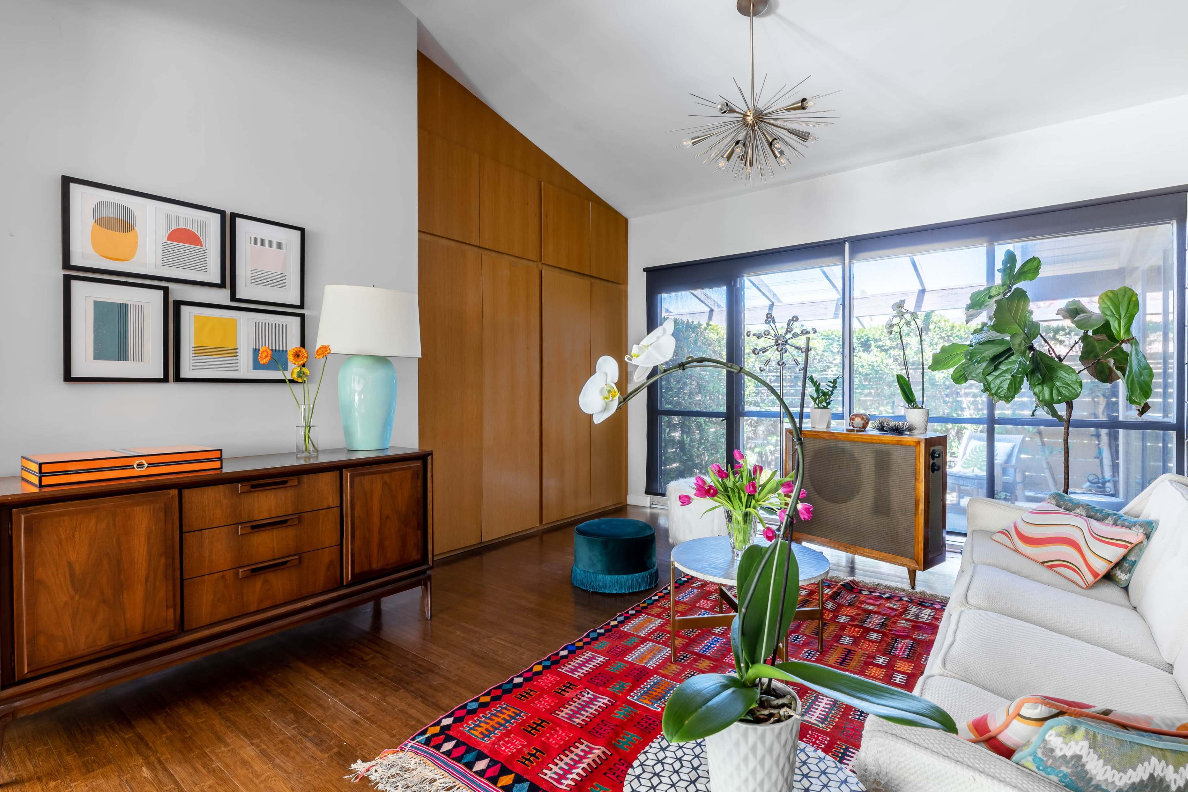 A living room features a mid-century modern sideboard, a large window with sliding doors, and a colorful rug under a bright white sectional sofa.