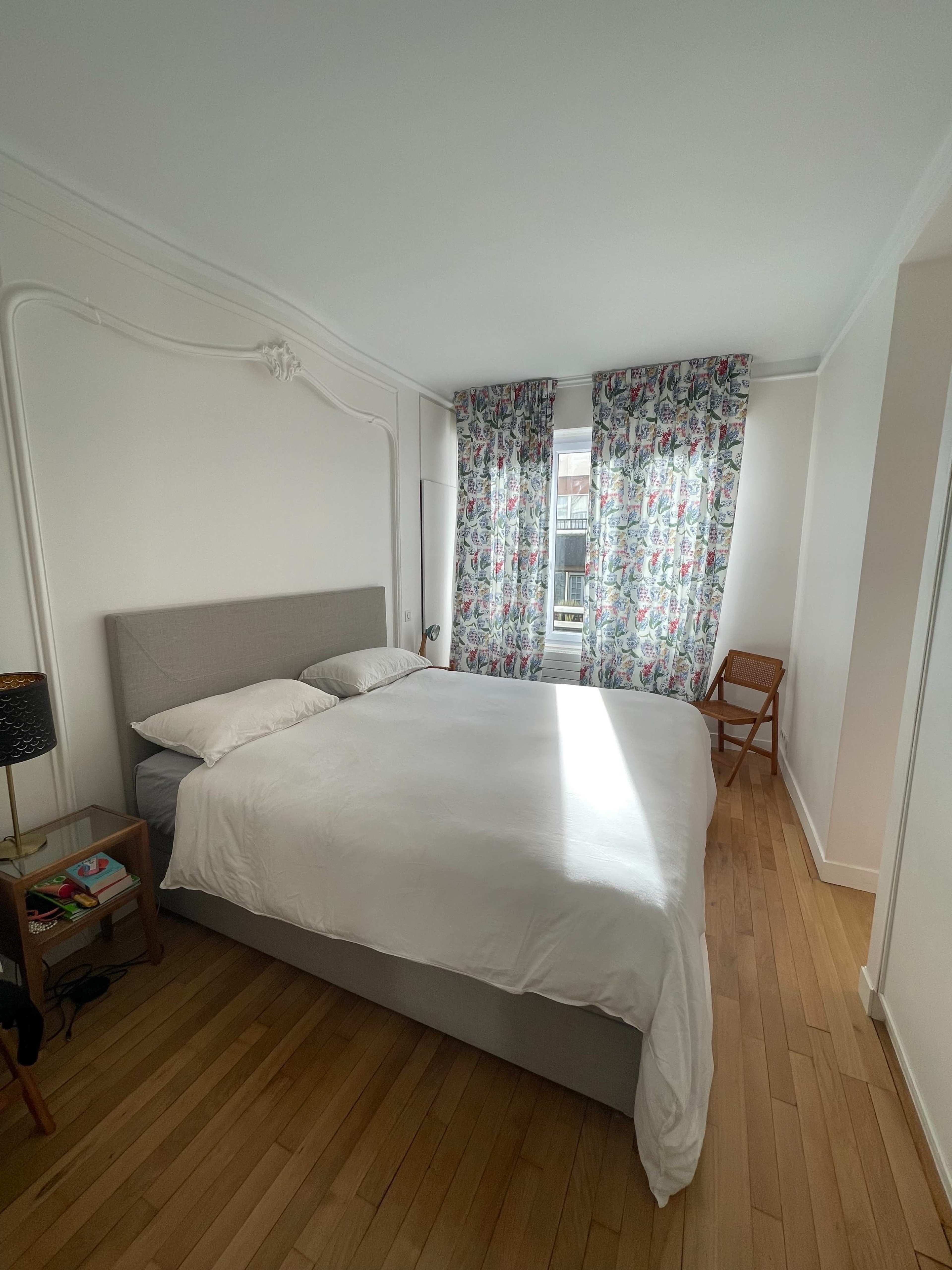 The image shows a simple bedroom with a large bed, floral patterned curtains, a wooden chair, and light wood flooring.