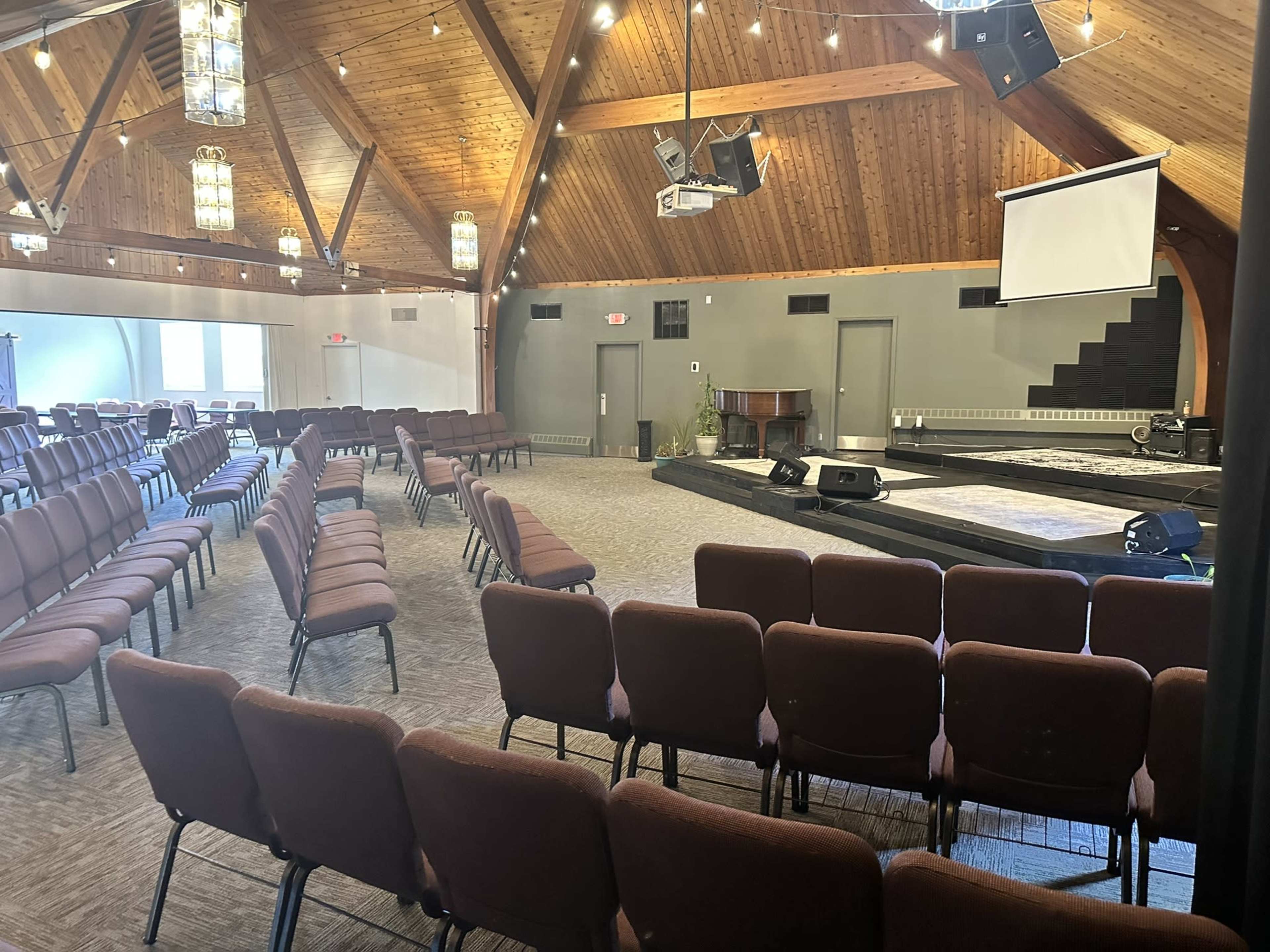 An empty indoor auditorium with wooden beams, rows of brown chairs facing a stage, and a large screen on the wall.