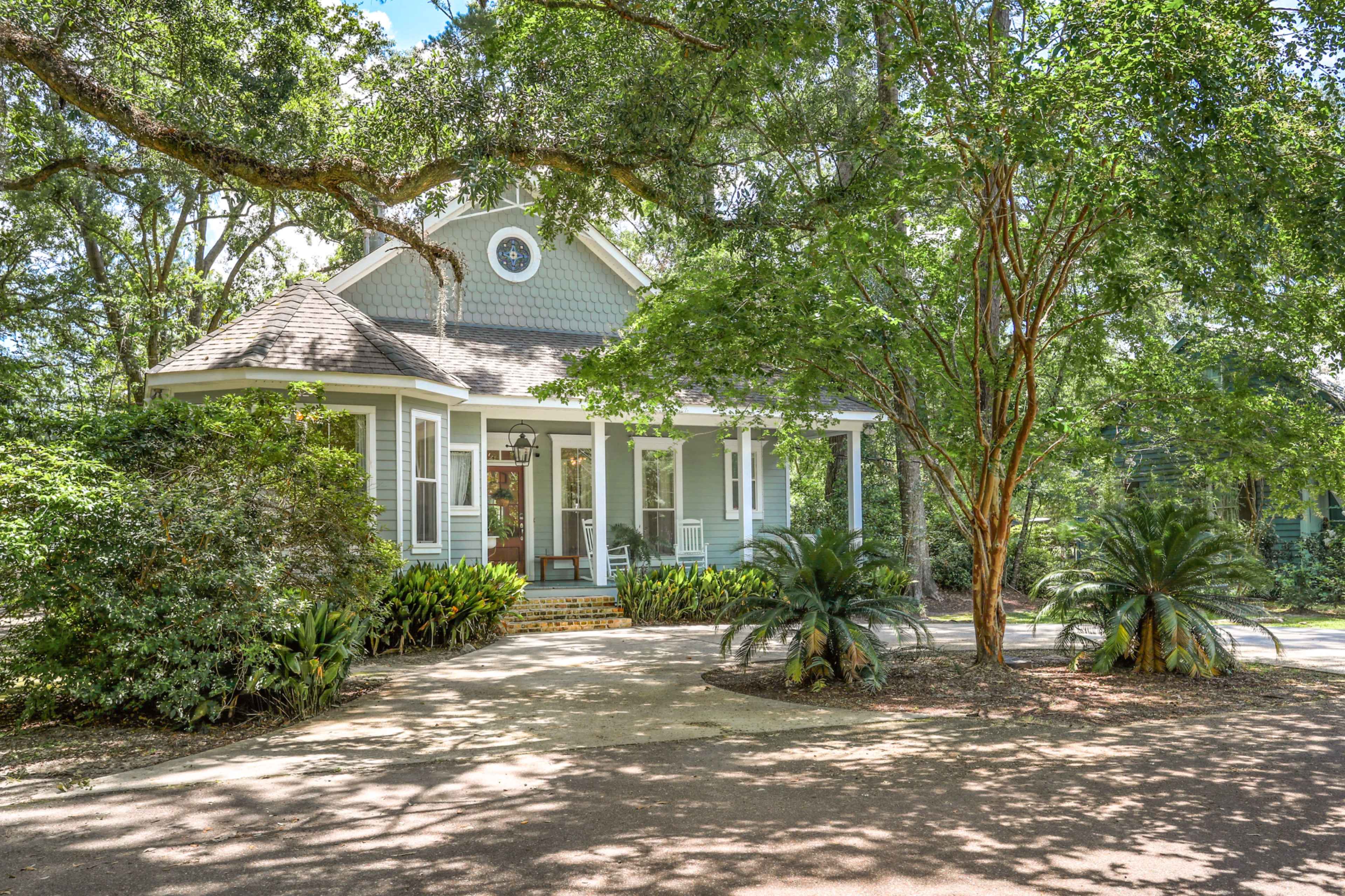 A light blue, two-story house is surrounded by lush greenery and trees, with a winding driveway leading up to the entrance.