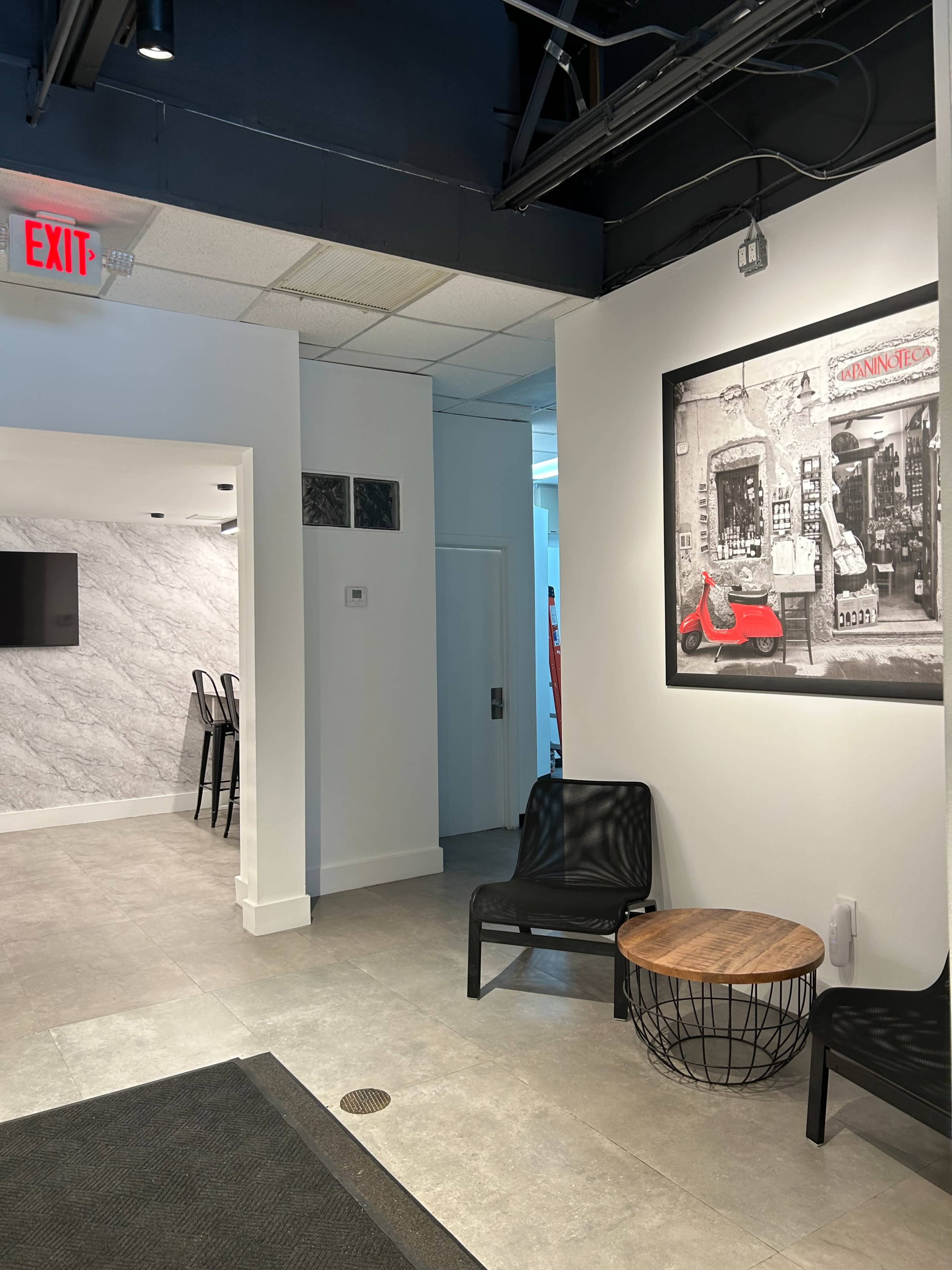 The image shows a modern interior space featuring a seating area with black chairs and a round wooden table, alongside a wall with a framed black-and-white photograph of a storefront.