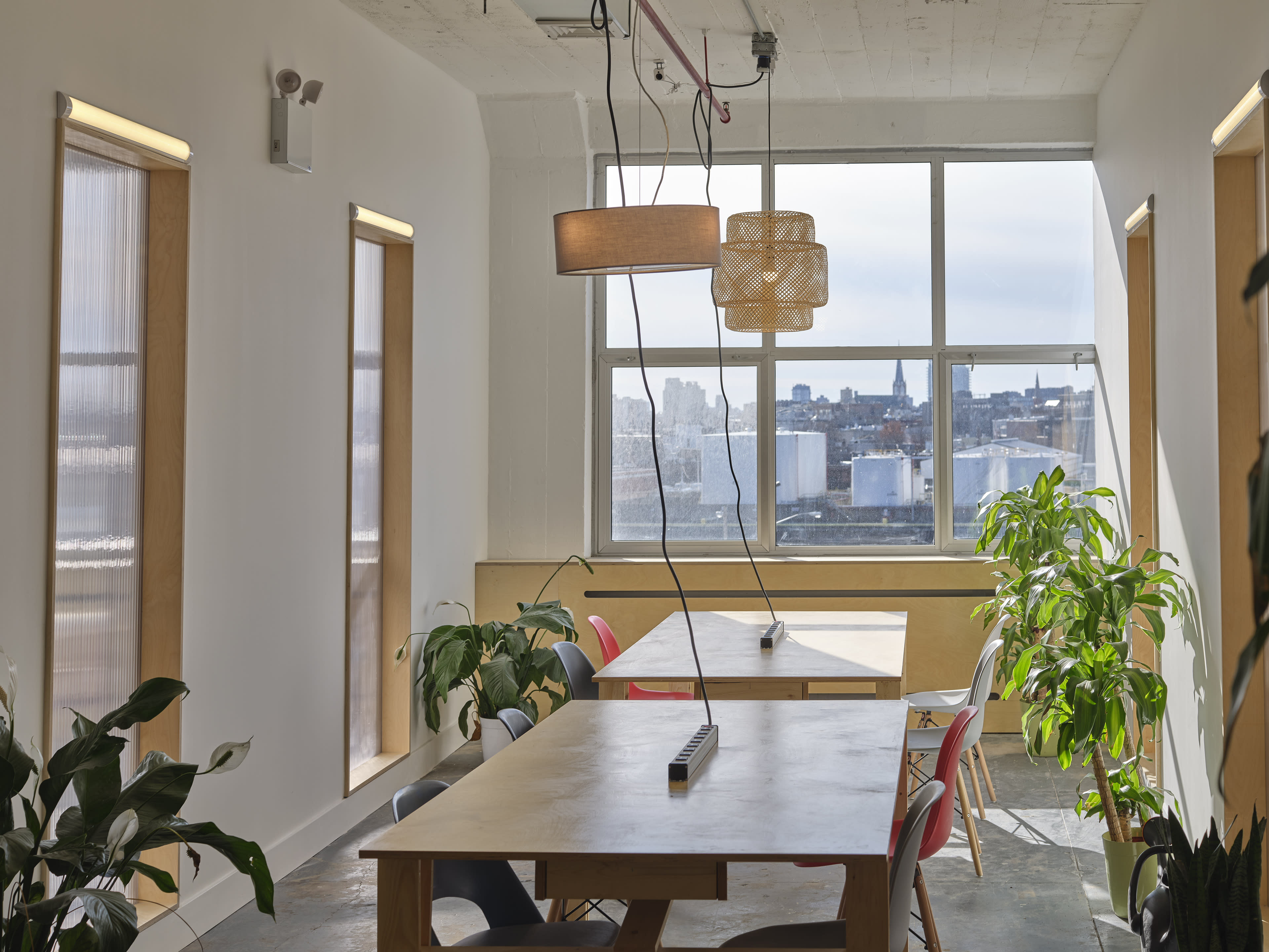 A bright, minimalist office space with tables, plants, and large windows offering a view of the city.