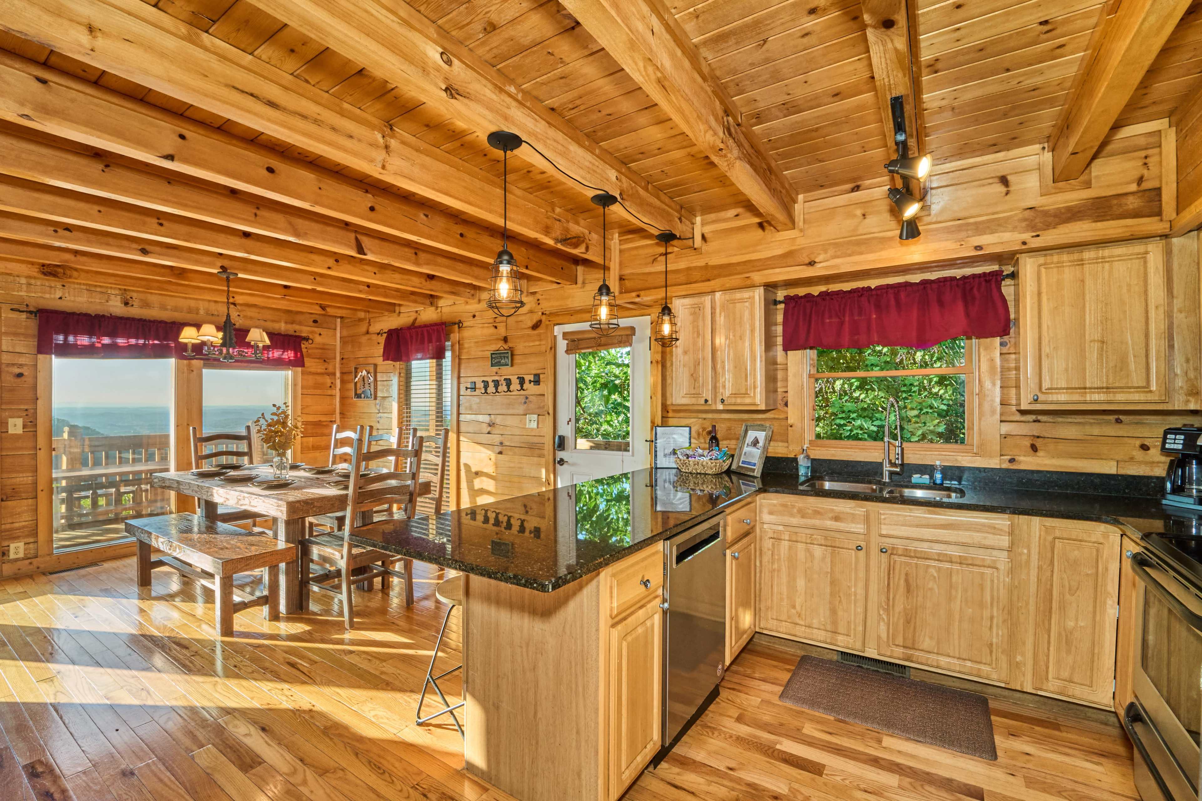 The image shows a wooden cabin kitchen with a dining area, featuring a black countertop, wooden cabinets, and a view of nature through large windows.