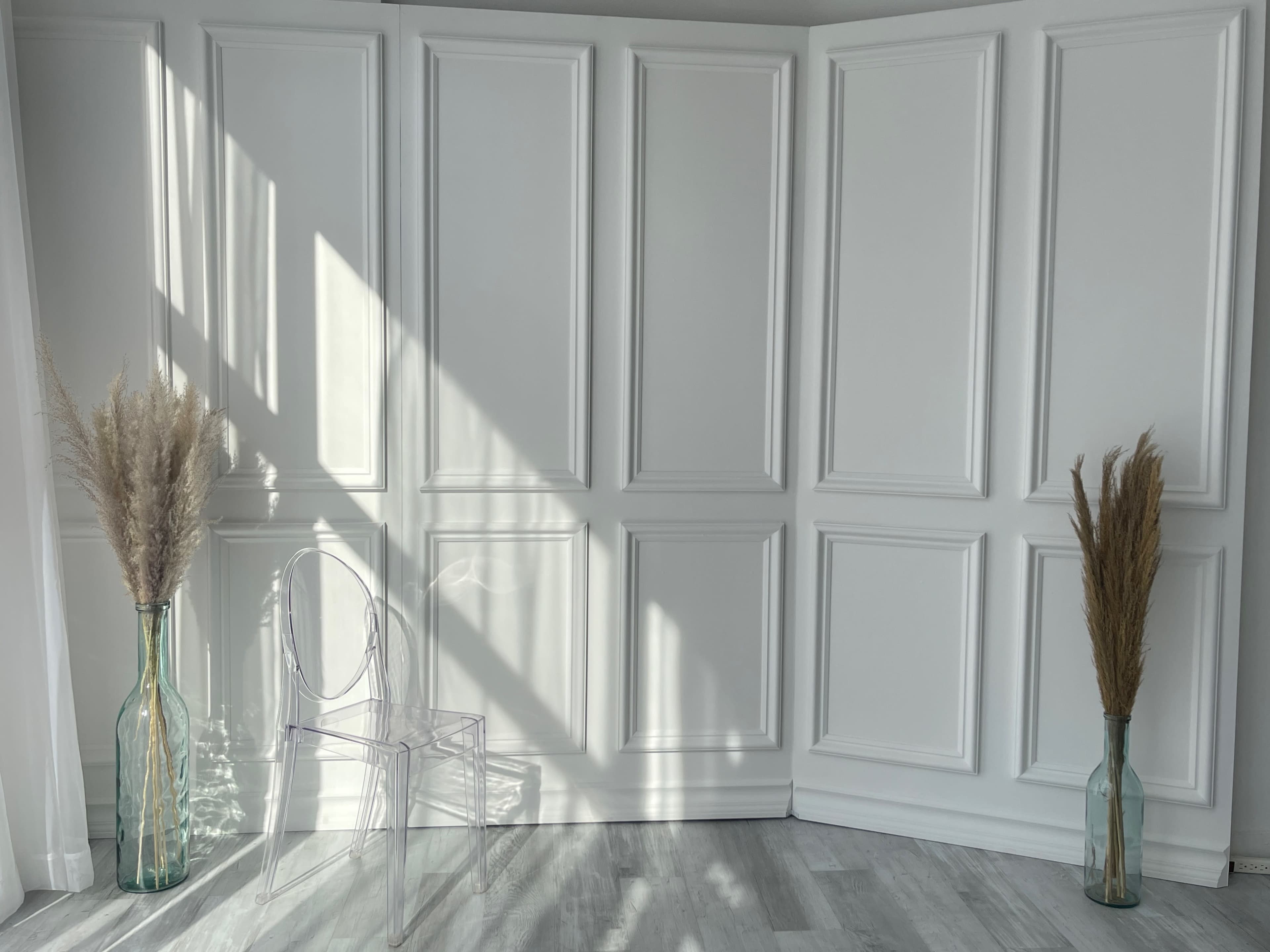 A minimalist interior with a clear acrylic chair and two vases containing dried plants, positioned against a backdrop of white-paneled walls illuminated by sunlight.