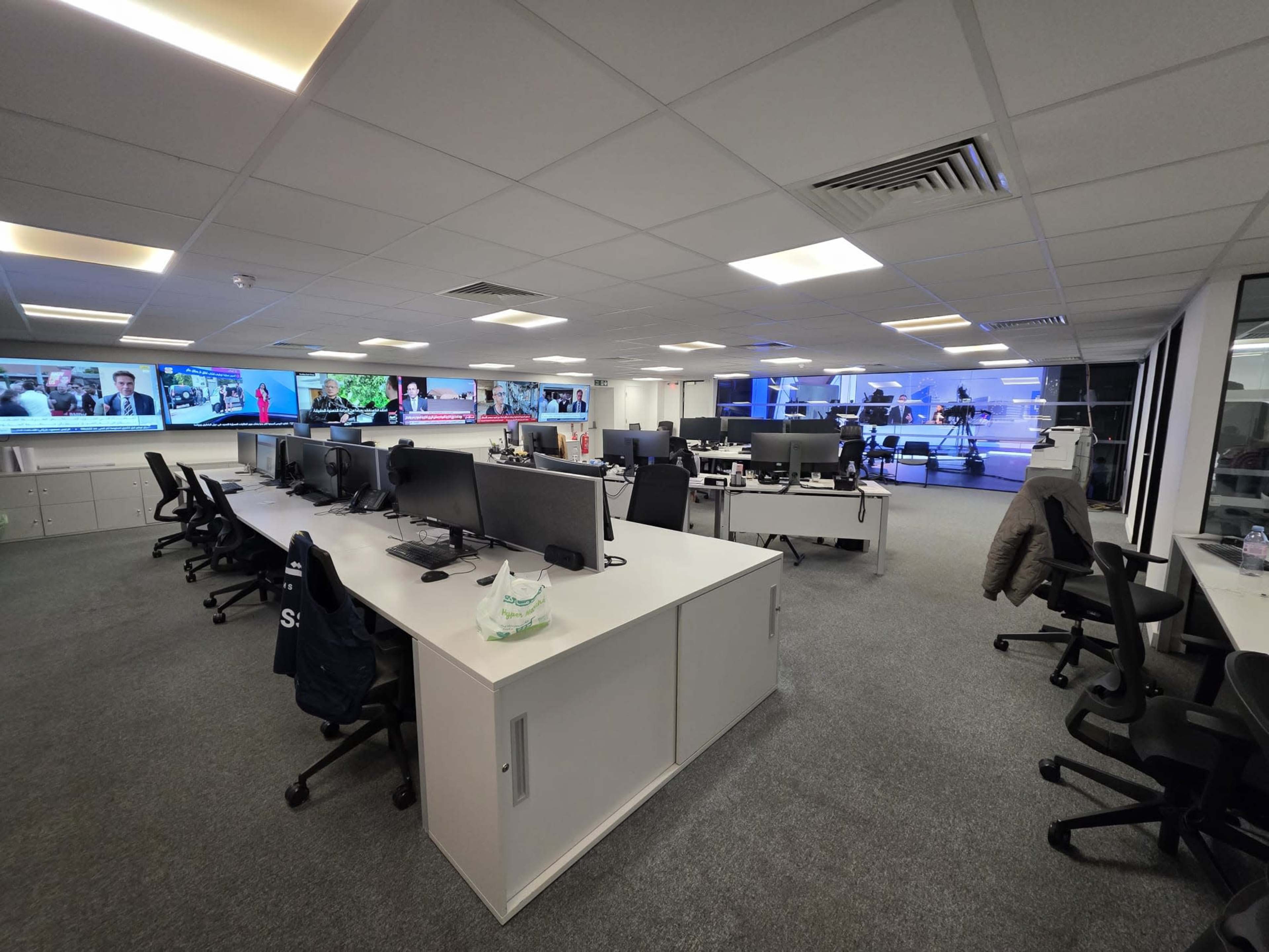 The image shows a modern, empty office space with several desks and computer monitors, and large screens displaying news broadcasts on the walls.