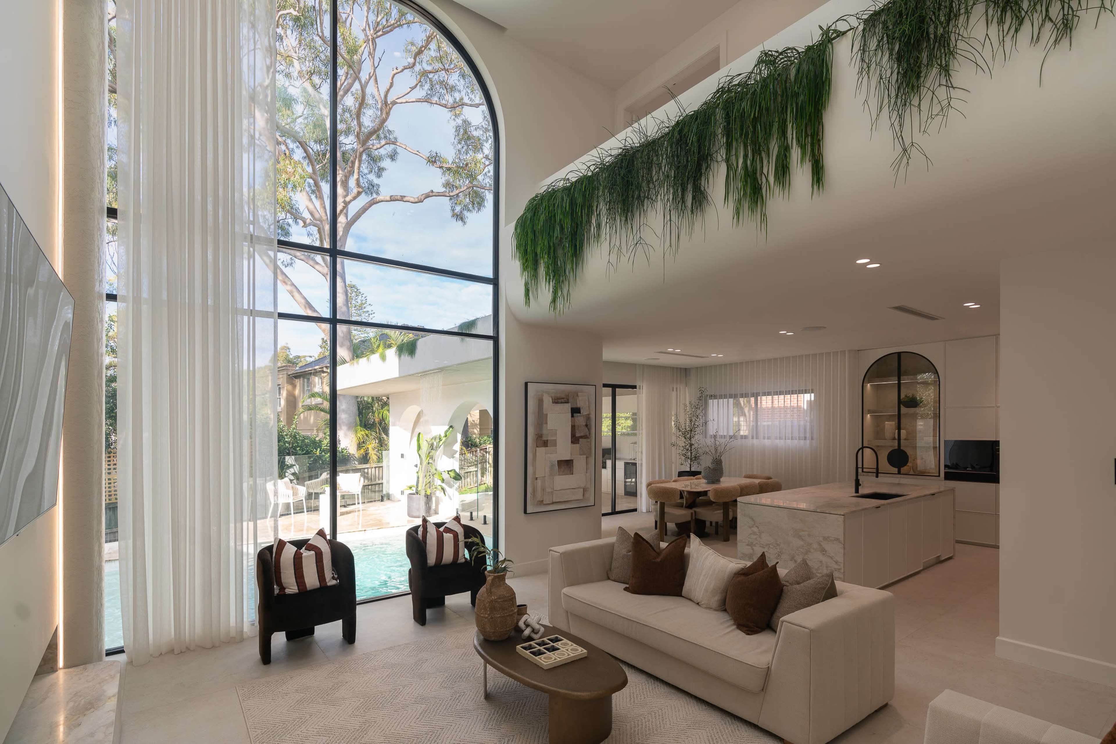 The image shows a spacious, modern living area with large windows, a light color palette, and contemporary furniture, looking out to a pool area.