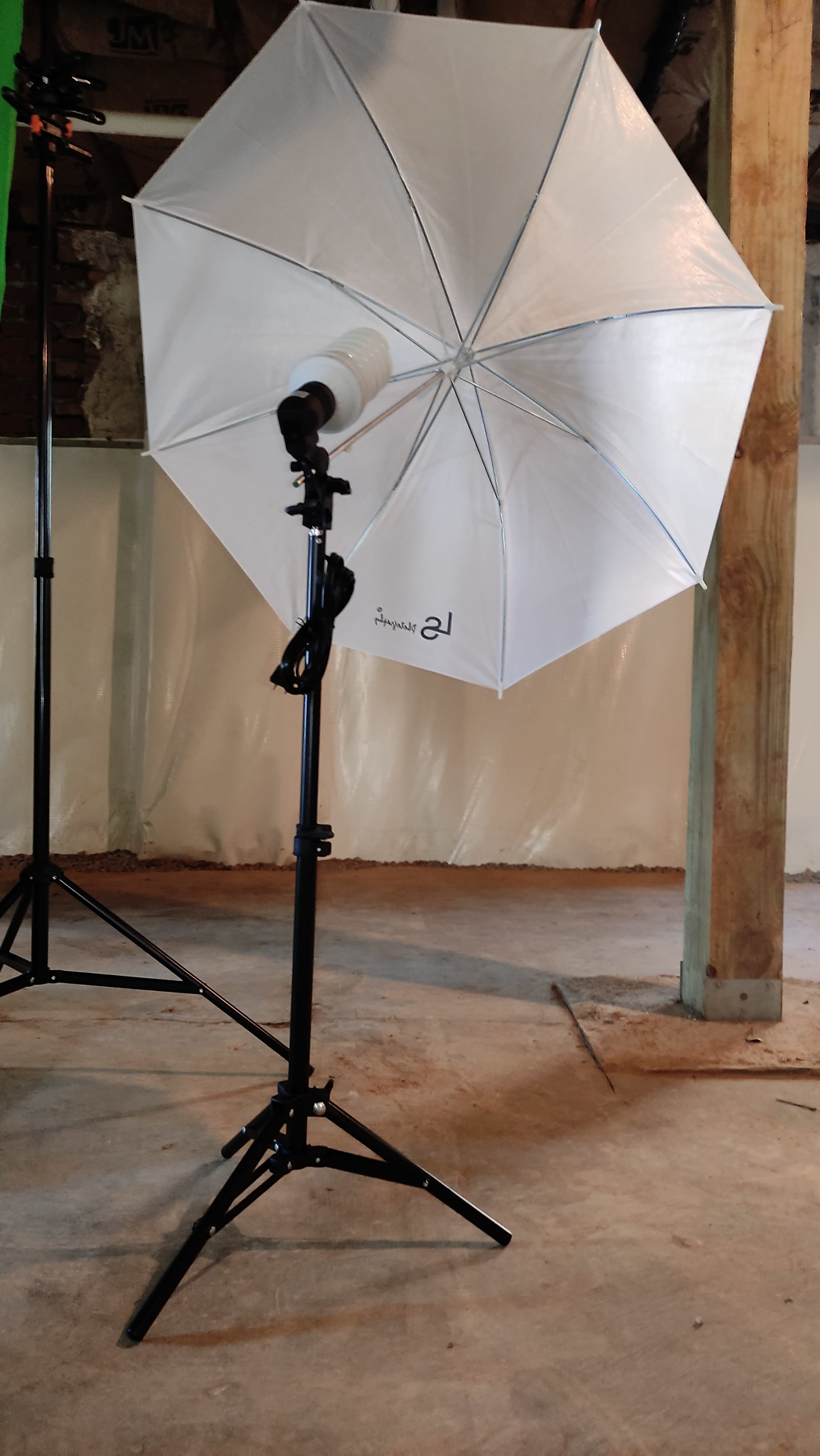 A photography light umbrella is positioned on a stand in an unfinished room with concrete flooring and exposed wooden beams.