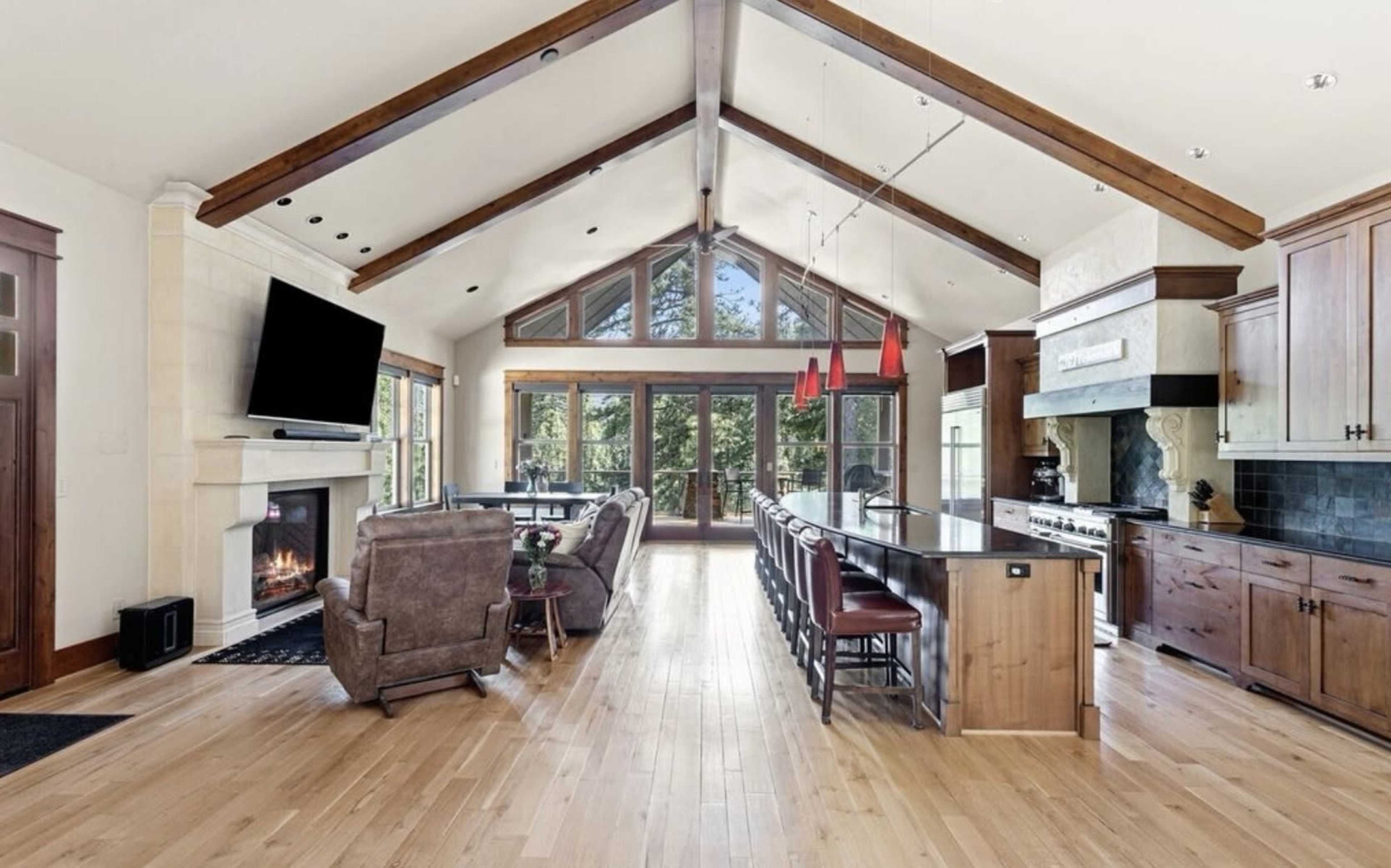 A spacious kitchen and living area feature high wooden beams, a large fireplace, and a long kitchen island with bar seating.