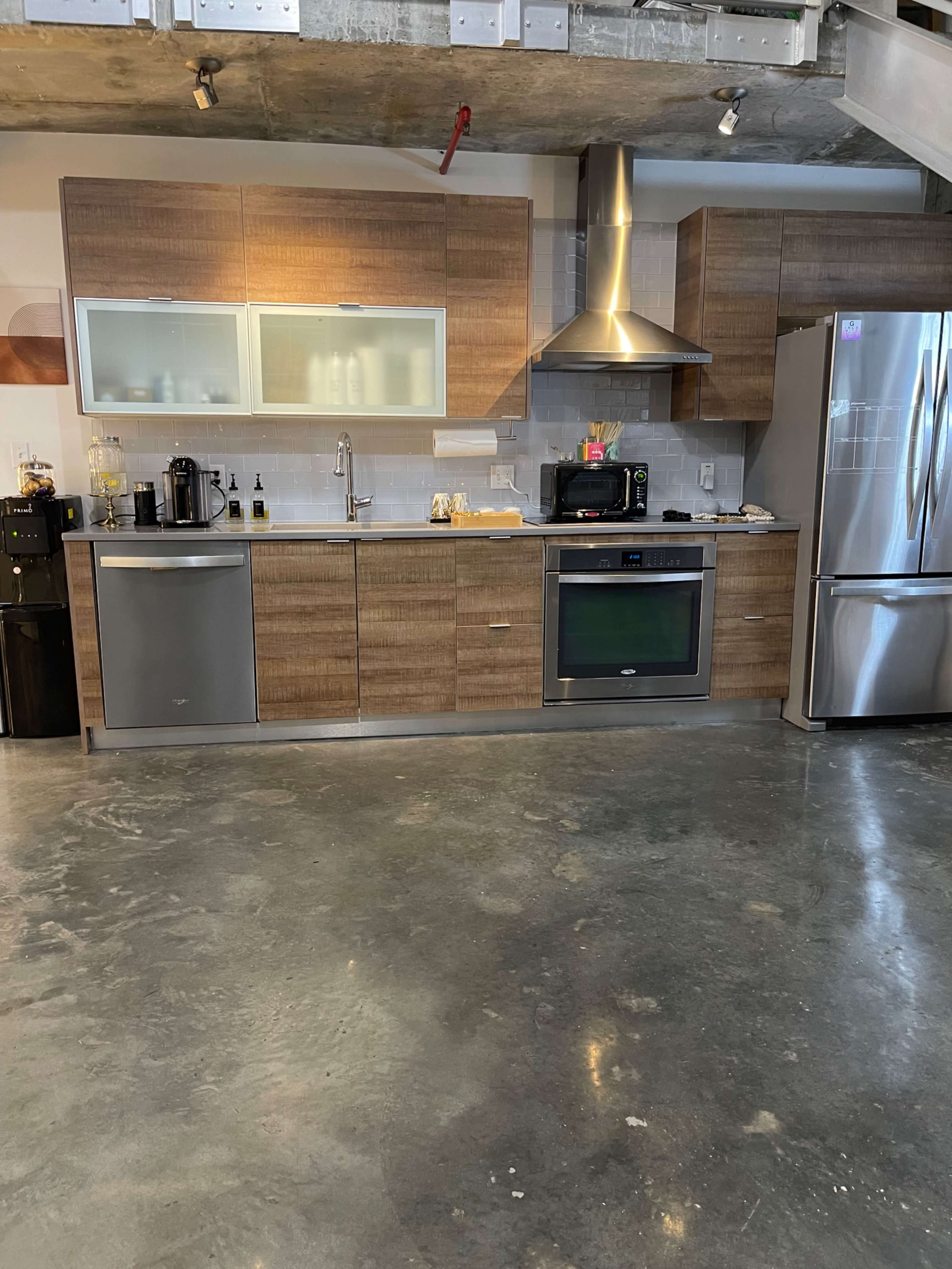 The image shows a modern kitchen with wooden cabinets, stainless steel appliances, and a concrete floor.