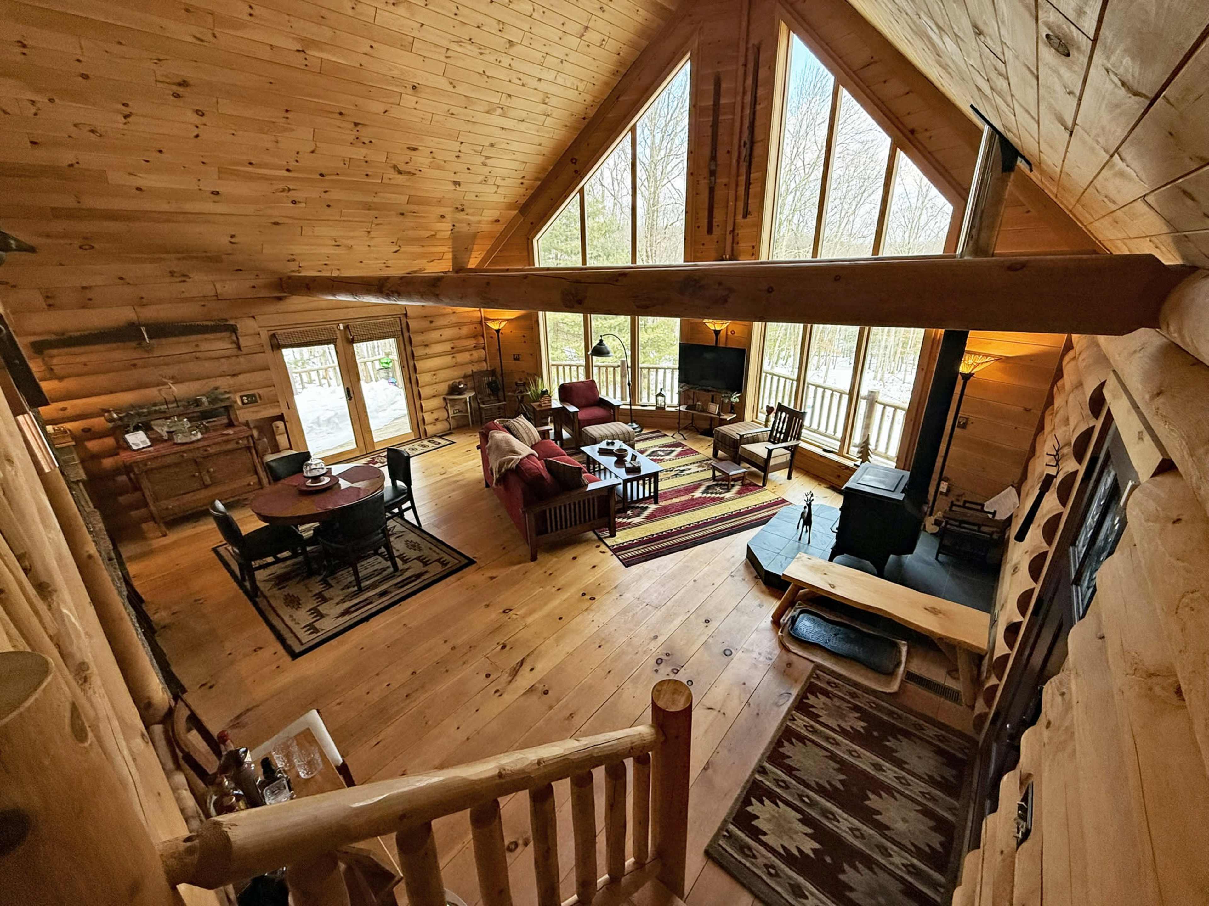 The image shows a rustic wooden cabin interior with a high ceiling, large windows, a living area featuring comfortable seating, and a dining table.