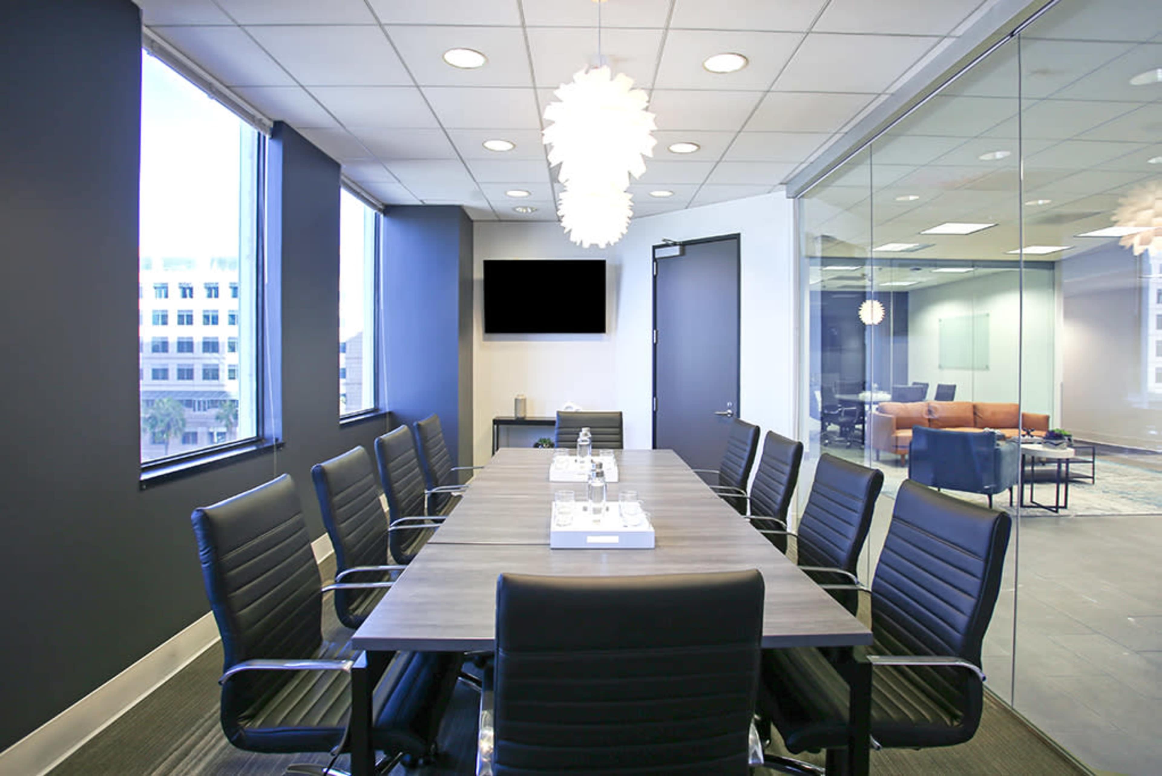 A modern conference room features a long table surrounded by black chairs, with windows providing natural light and a wall-mounted television.
