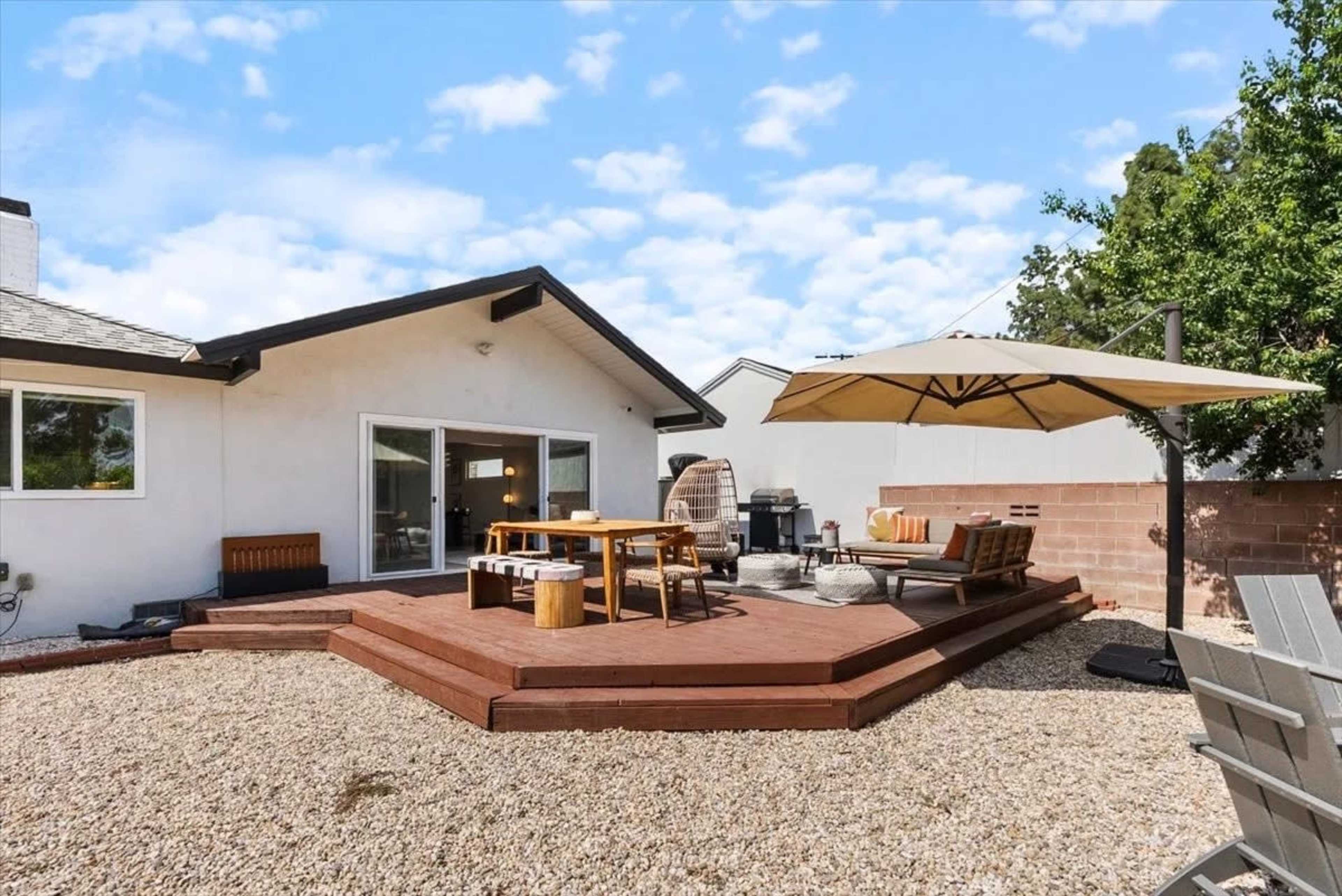 The image depicts a backyard with a wooden deck, outdoor furniture, and an umbrella, surrounded by gravel and a low wall.