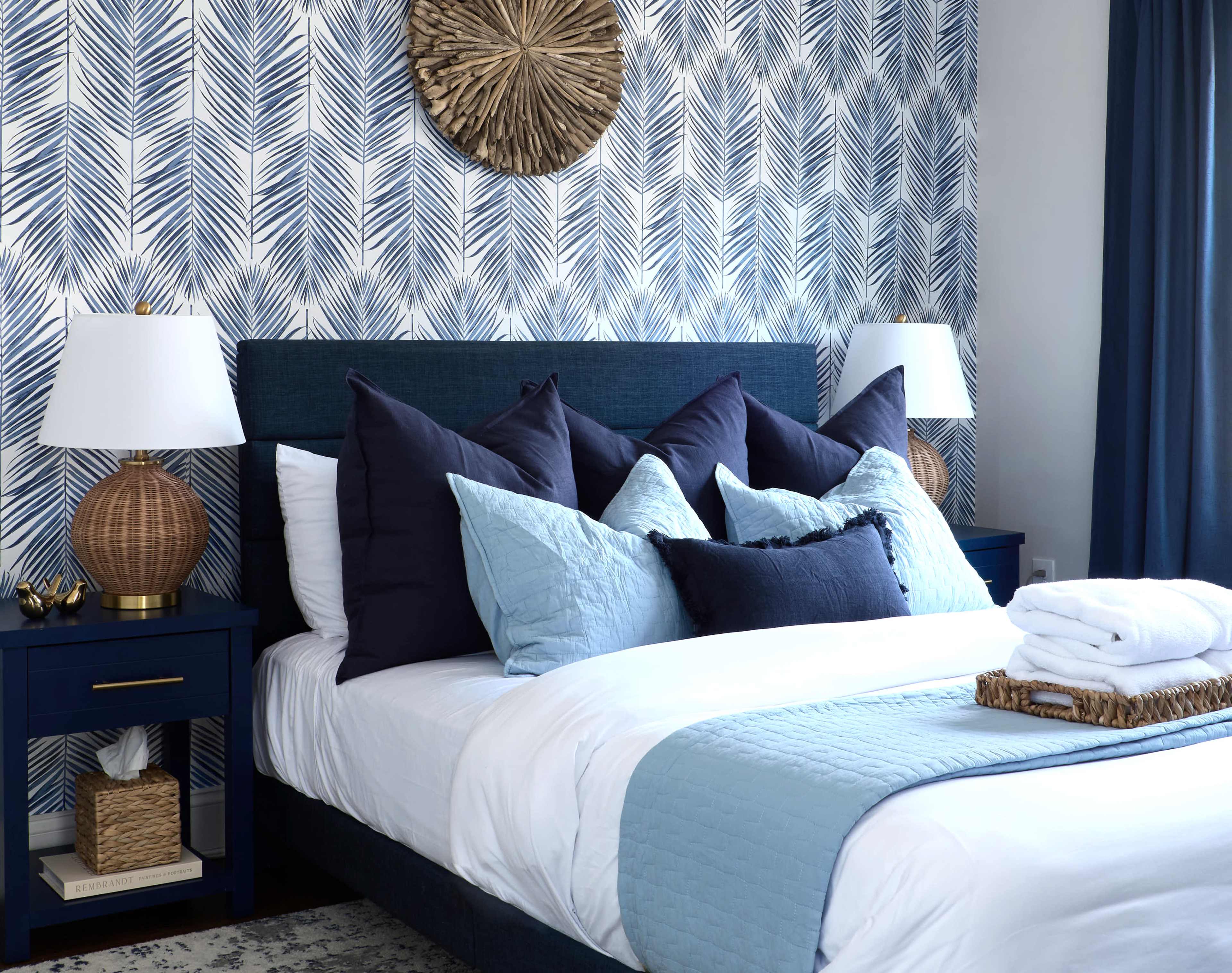The image shows a neatly arranged bedroom featuring a dark blue bed with multiple pillows, two bedside tables with lamps, and a patterned wallpaper in shades of blue and white.