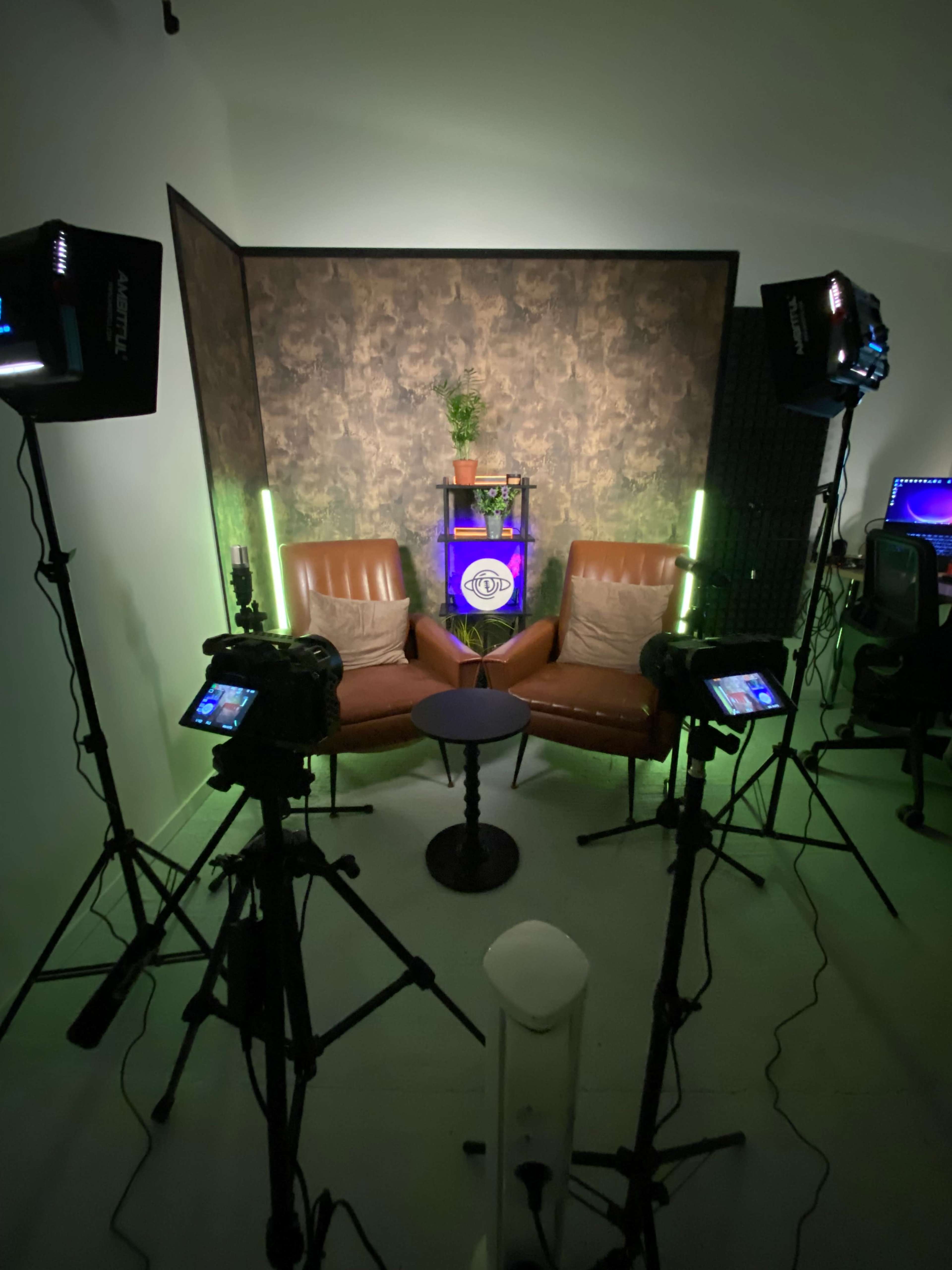 The image shows a small, well-lit studio setup featuring two brown chairs facing each other, a small round table in between, and various lighting equipment positioned around the scene.