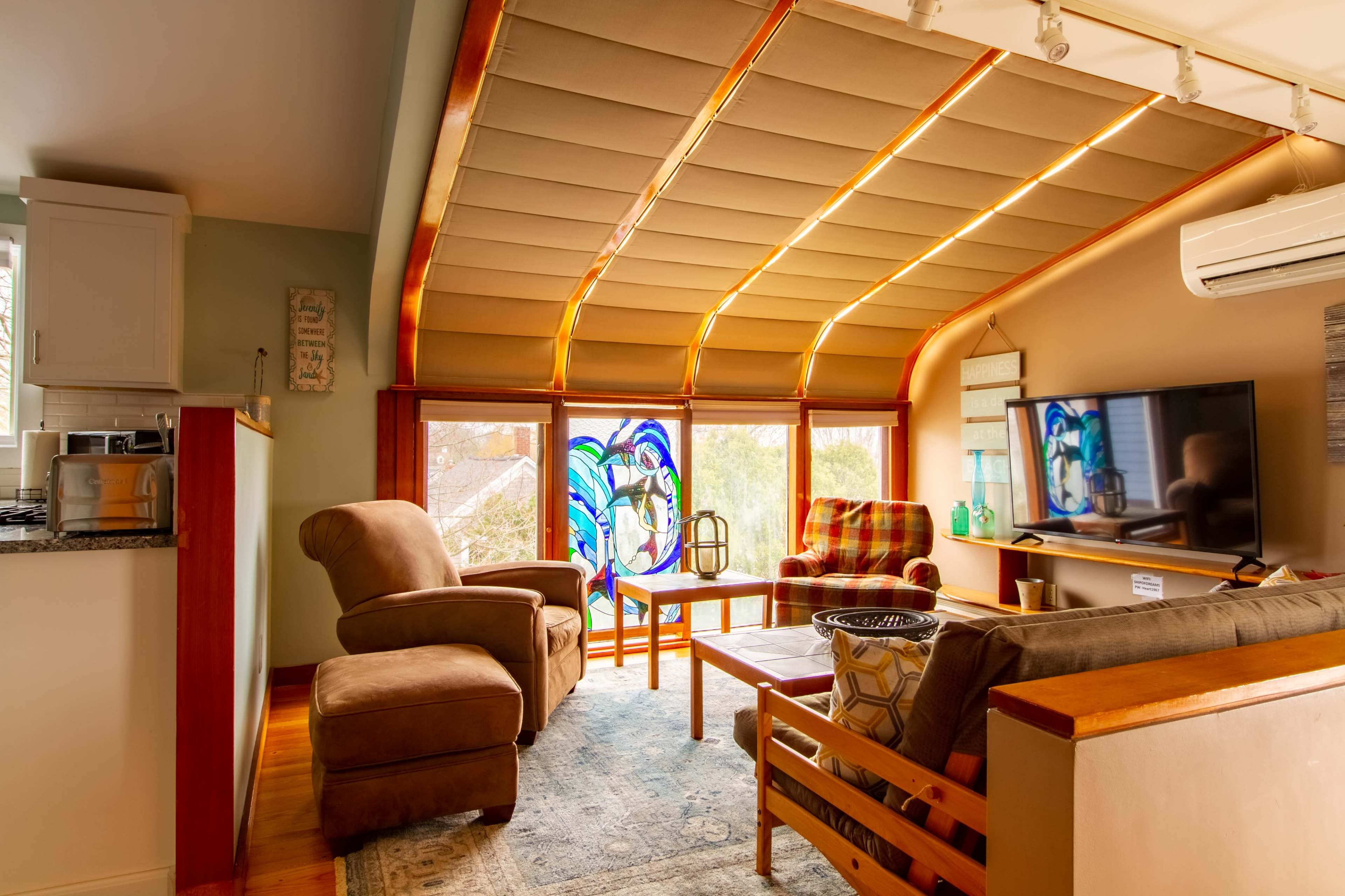 The scene features a cozy living area with a combination of seating, a large window adorned with stained glass, and a television mounted on the wall.
